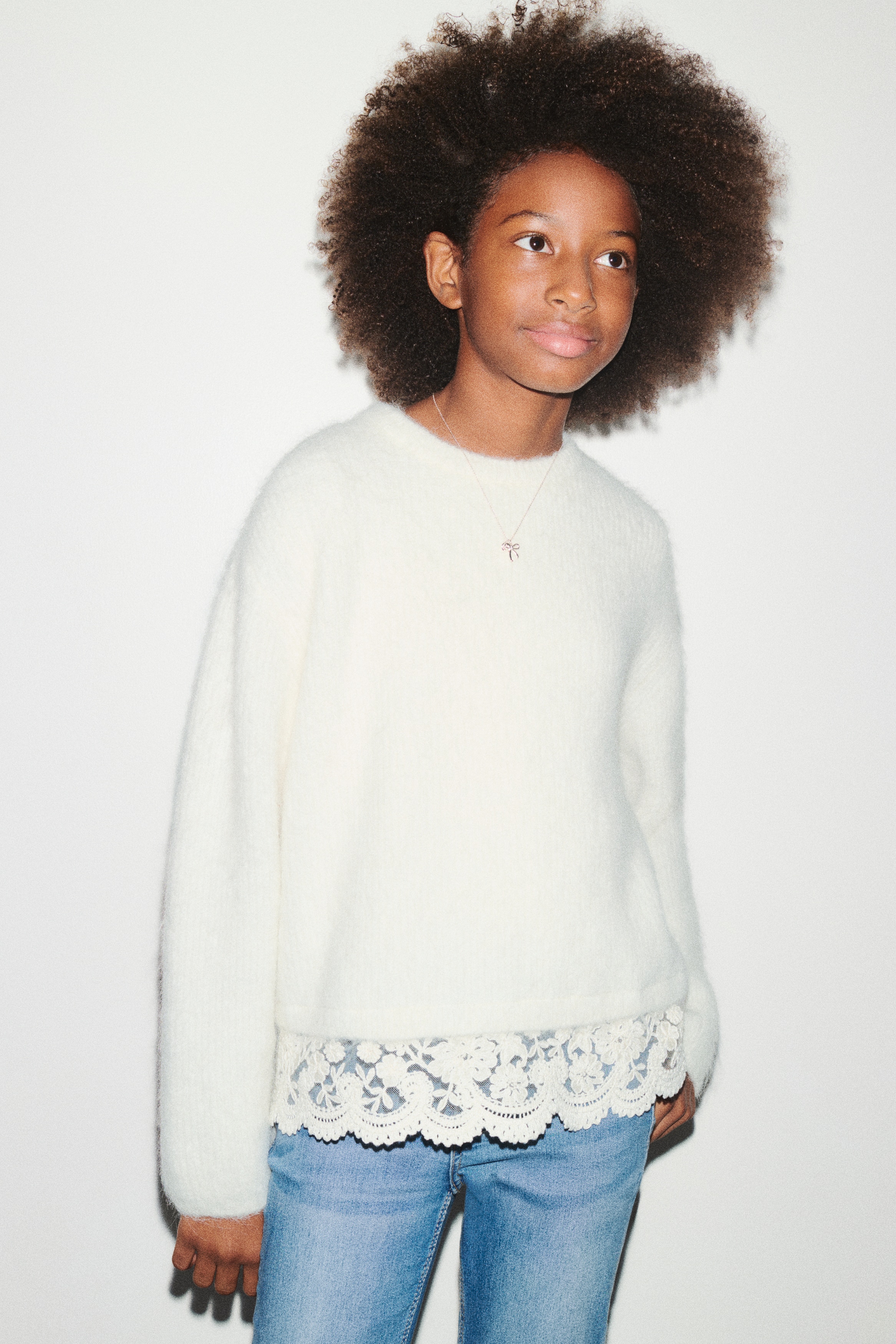 A cream-coloured knit jumper, facing front, features a soft, fuzzy texture with a ribbed crew neck and long sleeves. The garment is displaying a delicate, scalloped floral lace trim along its bottom hem.