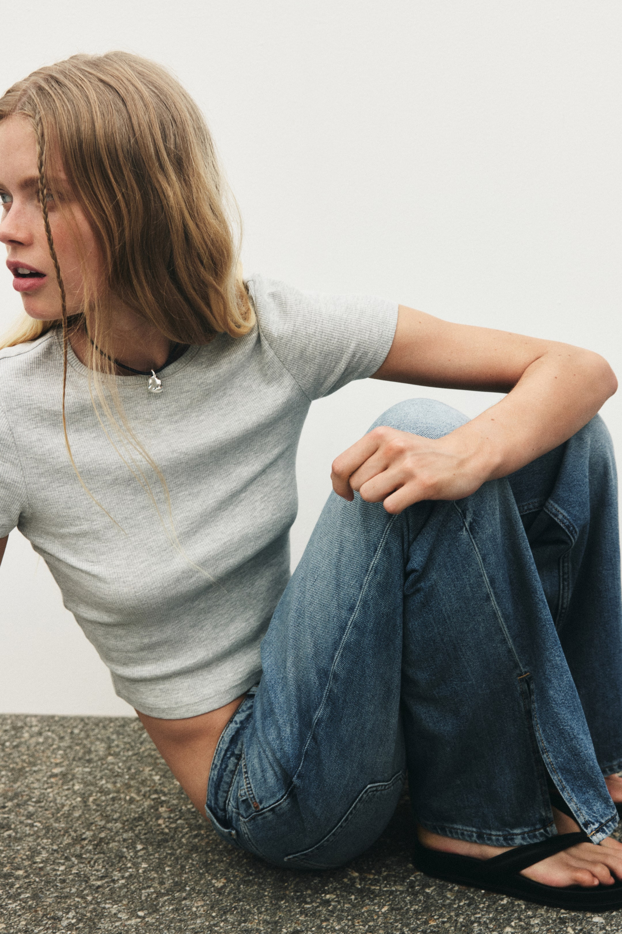 View larger image: The grey ribbed T-shirt, featuring short sleeves and a crew neck, is worn by the model. Its cropped hem is visible, styled with blue jeans and a black cord necklace.