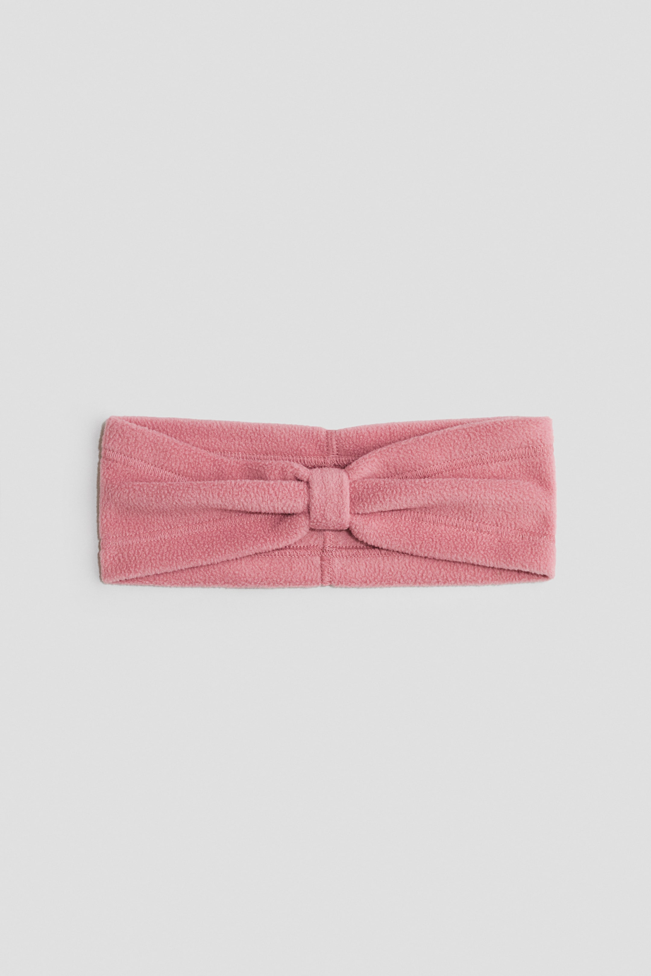 Grösseres Bild ansehen: Ein rosa Fleece-Stirnband liegt flach und ist mit einem zentralen Knoten-Detail versehen.