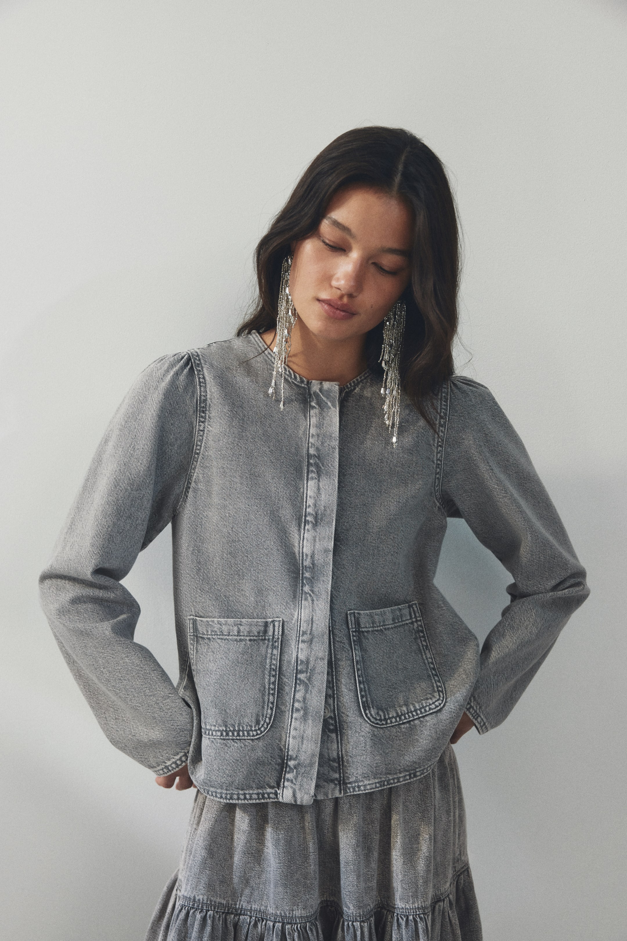 Grösseres Bild ansehen: Eine grau gewaschene Jeansjacke wird von vorne präsentiert, mit einem kragenlosen, runden Ausschnitt, einer verdeckten Knopfleiste vorne und langen Ärmeln mit dezenten Puffschultern. Zwei große, abgerundete aufgesetzte Taschen sind auf der Vorderseite sichtbar, und sie wird mit einem passenden grau gewaschenen Jeansrock mit Stufenoptik kombiniert.