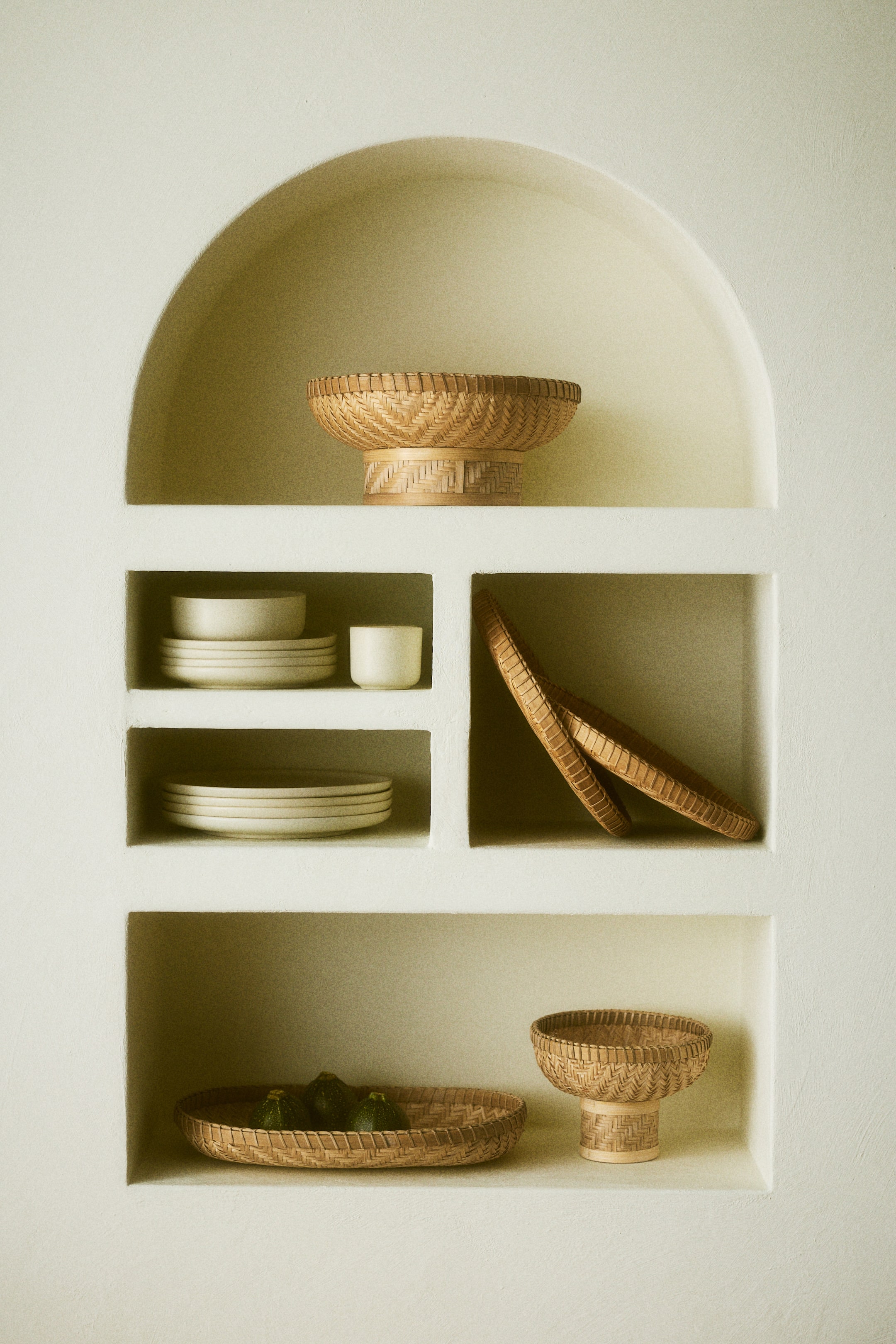 הצגת תמונה גדולה יותר: אוסף של סלים קלועים וכלי קרמיקה מוצג. שני קערות סלים קלועות מורכבות מונחות על בסיסי פדסטל, וסל קלוע אובלי מחזיק פירות ירוקים. ערימות של צלחות קרמיקה בצבע בהיר וכוס מוצגות גם הן, עם שני מגשים קלועים עגולים שנשענים.