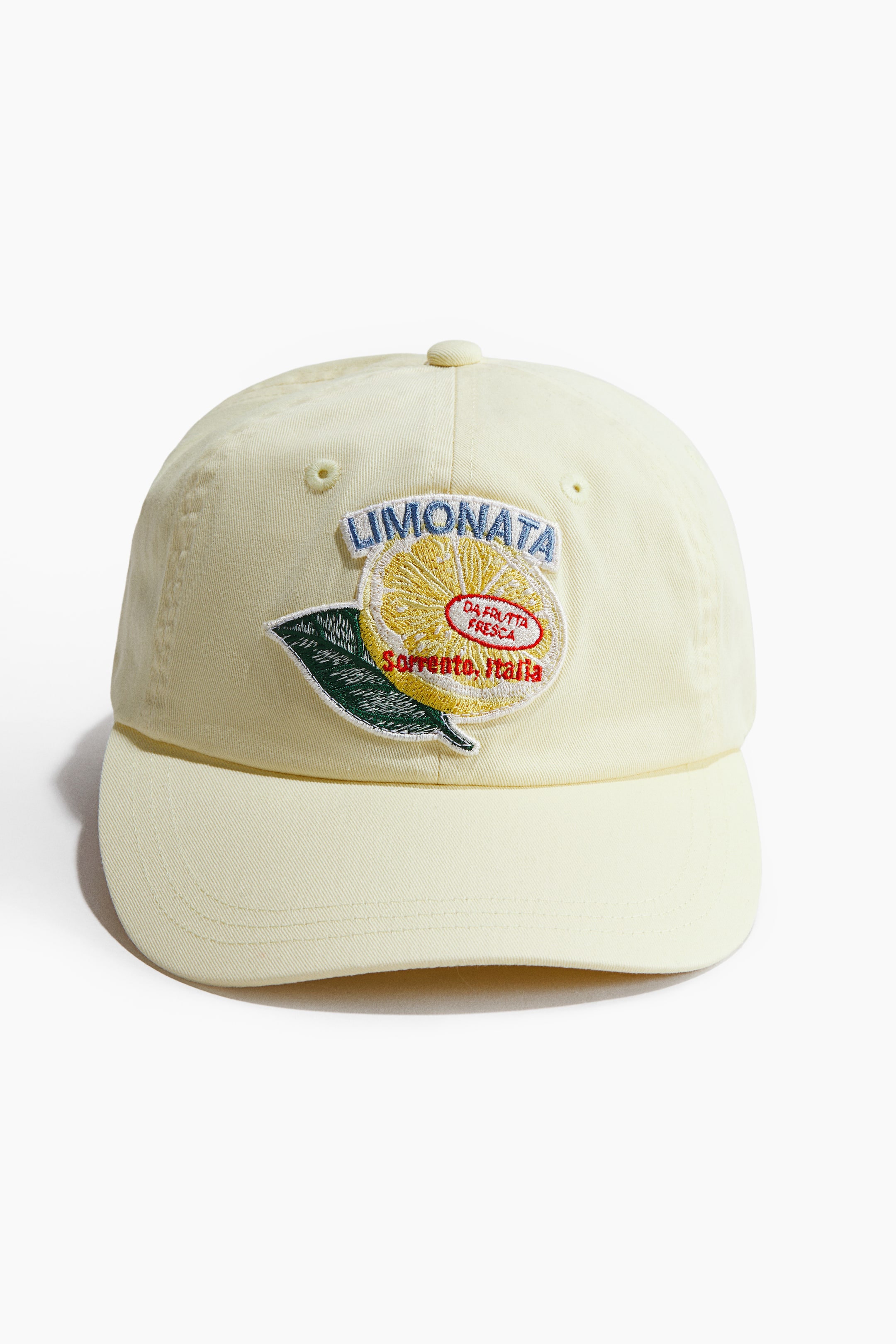 View larger image: A light yellow cotton twill cap is showcased from the front, highlighting an embroidered patch depicting a lemon slice with a green leaf and the word 'LIMONATA' in blue text above it.