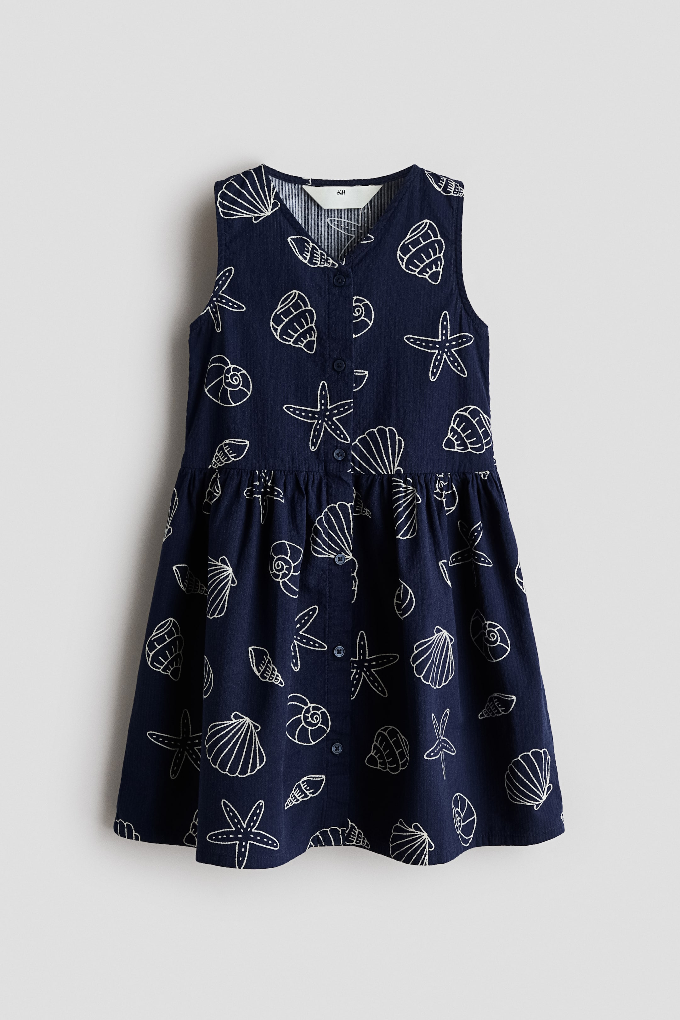 View larger image: "This sleeveless dress showcases a dark blue fabric adorned with a white seashell and starfish motif, as seen from the front. It features a V-neckline, a front button placket, a gathered waist seam, and a flared skirt."