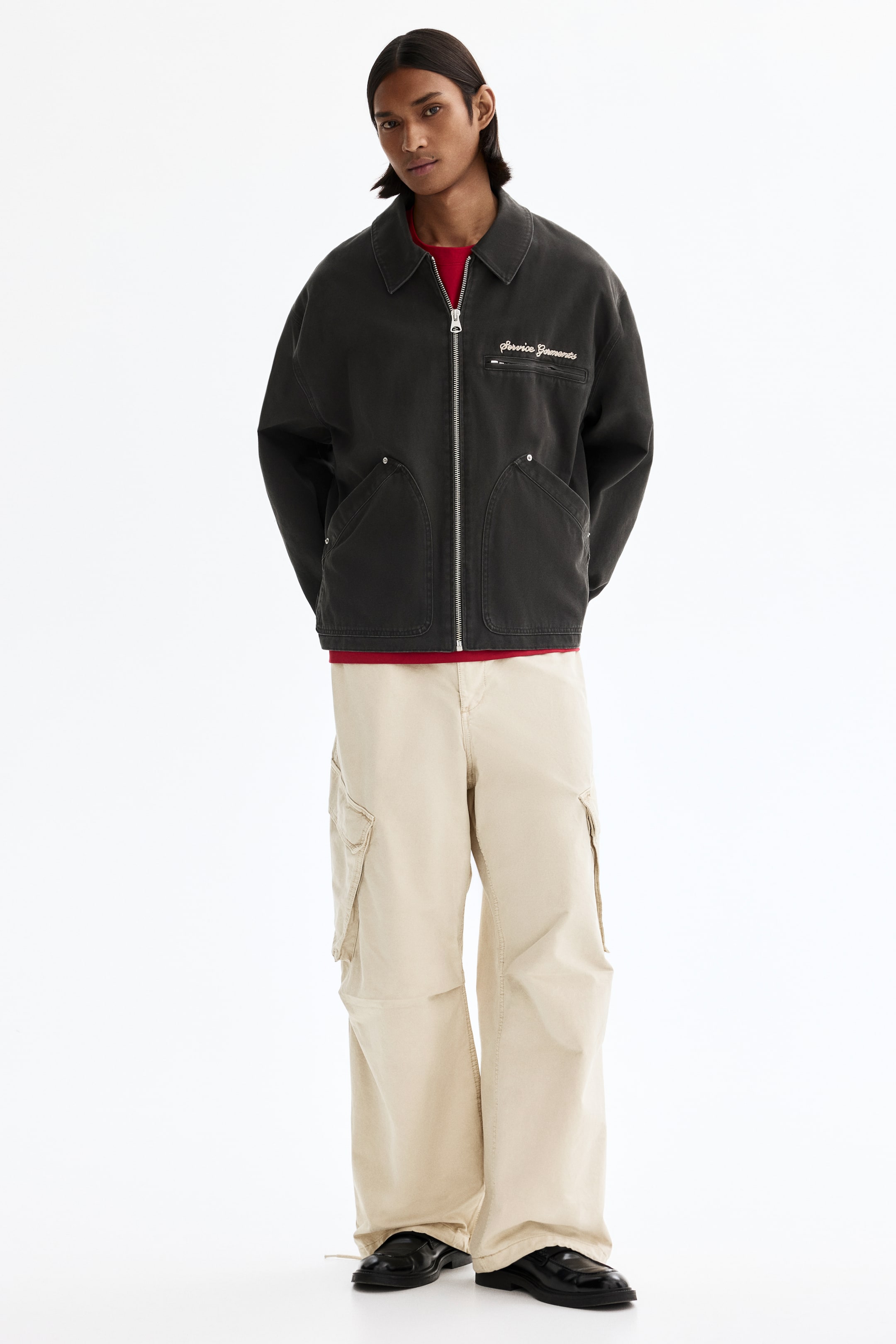 View larger image: A dark grey workwear jacket features a pointed collar, a full-length silver-toned zipper down the front, and two large rounded patch pockets with rivets. White embroidered text reading "Service Garments" is visible on the left chest, with a red T-shirt layered underneath.