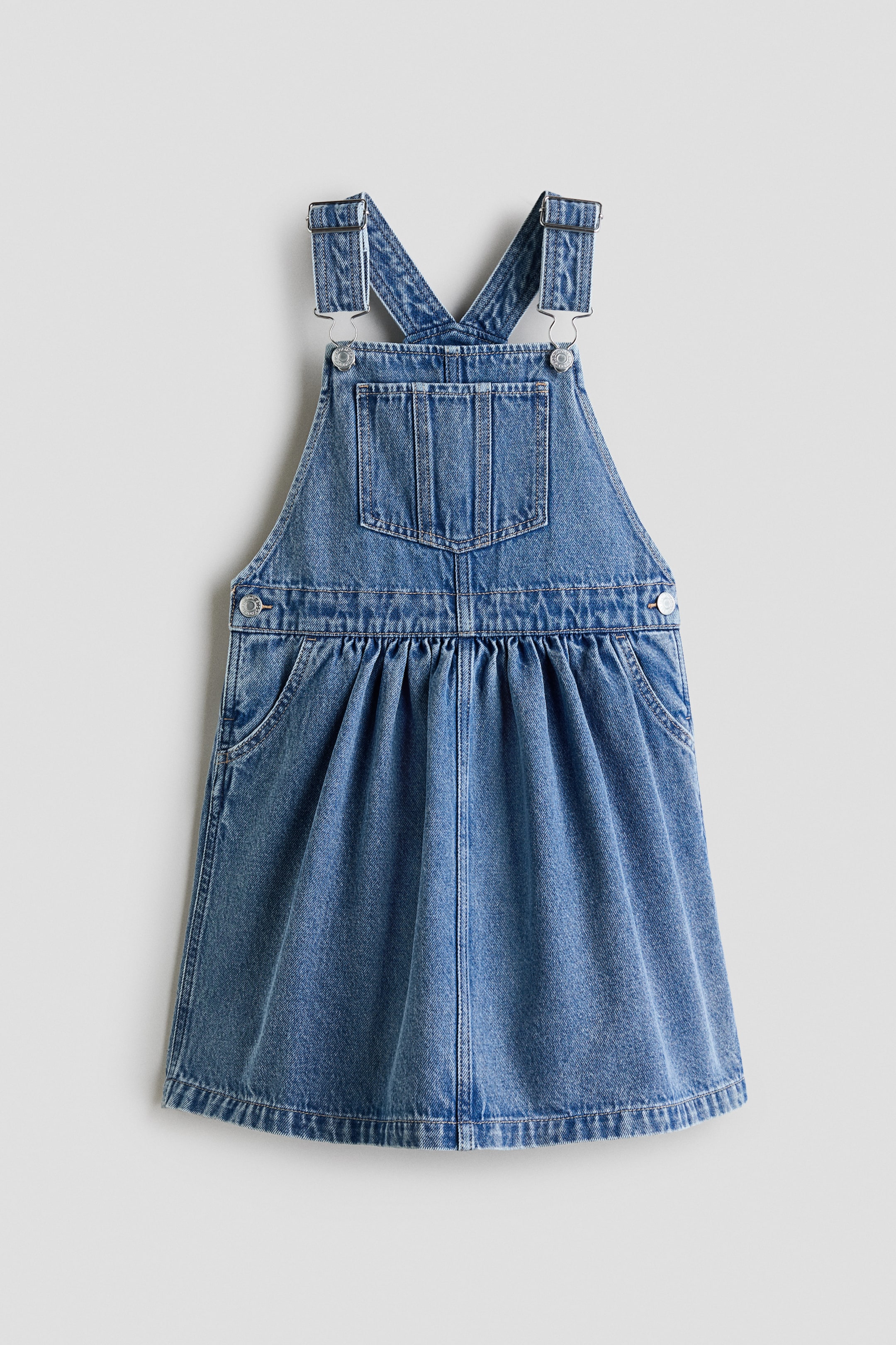View larger image: A light blue denim pinafore dress is displayed front-facing. It features adjustable shoulder straps with metal buckles and a square bib that includes a flap patch pocket. An elasticated waistline creates gathers, and two side pockets along with metal side buttons appear at the sides.