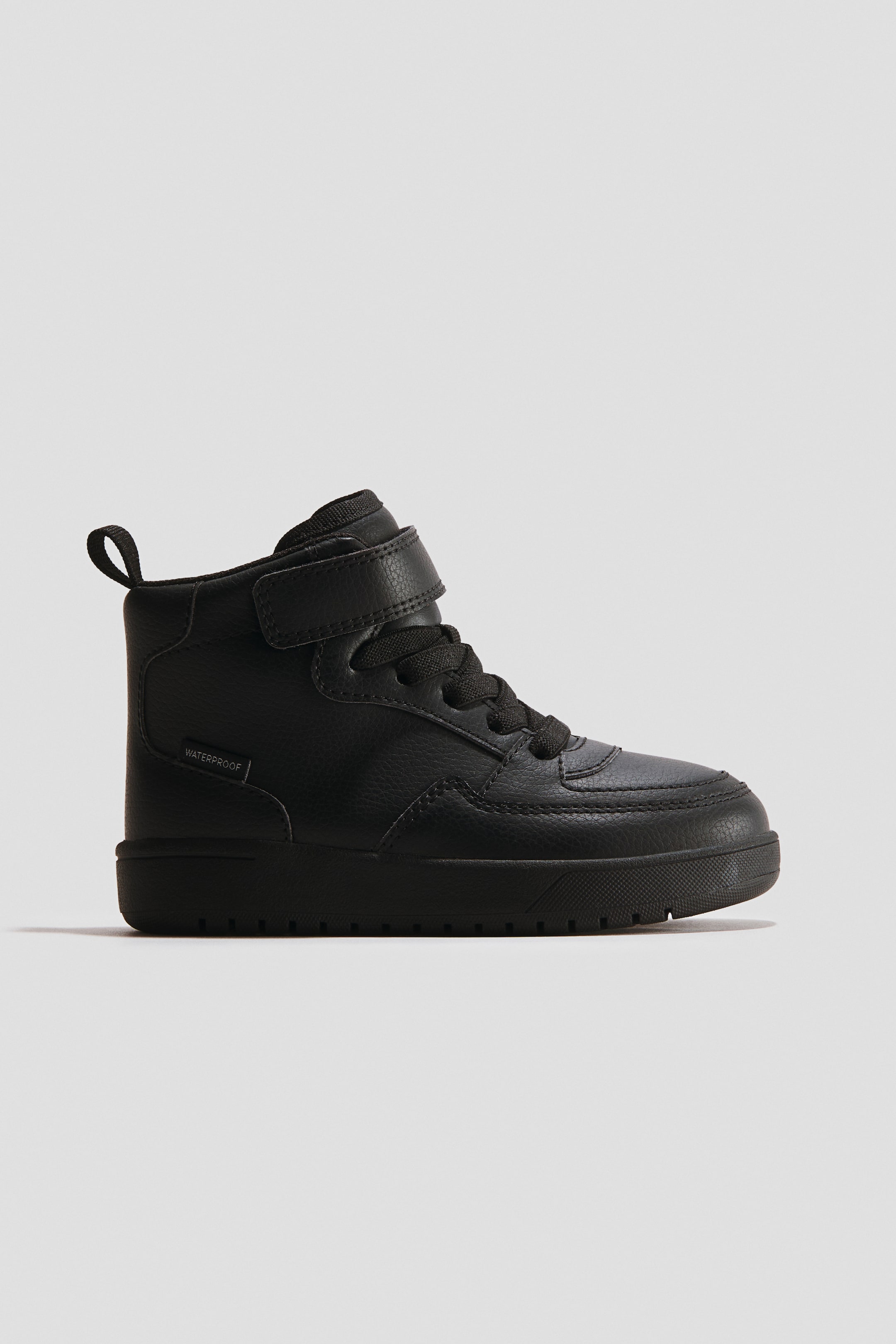 View larger image: A black high-top sneaker is displayed with a textured upper, black laces, and a single hook-and-loop ankle strap. It features a padded collar with a pull tab, a "WATERPROOF" label on the side, and a thick black sole with textured tread.