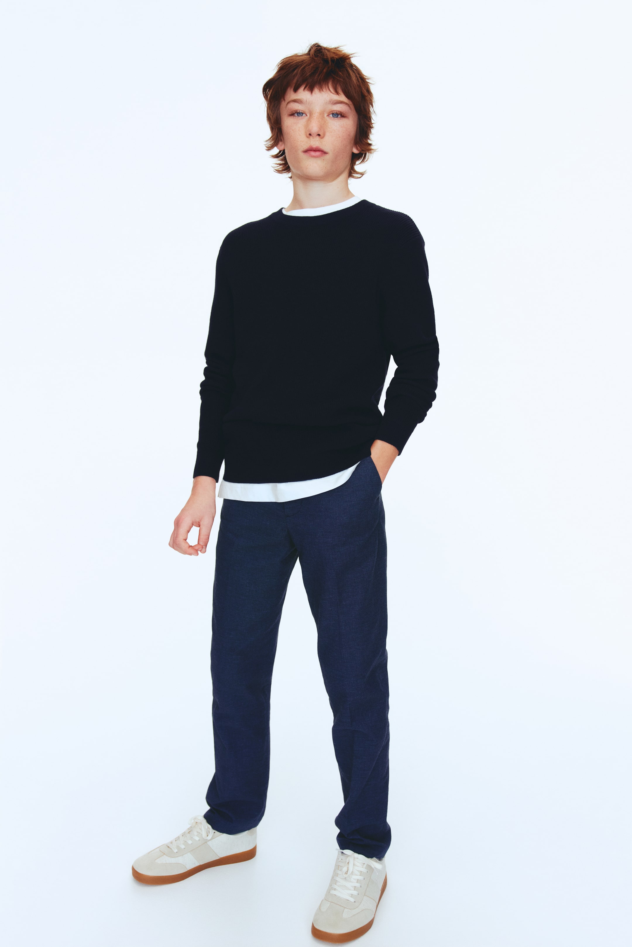 View larger image: A dark blue crew neck sweater, showcasing a ribbed hem, layered over a visible white t-shirt with its hem and neckline in view, is paired with dark blue textured trousers. The ensemble is finished with light-colored low-top sneakers featuring gum soles, all positioned forward to highlight the fit and design.