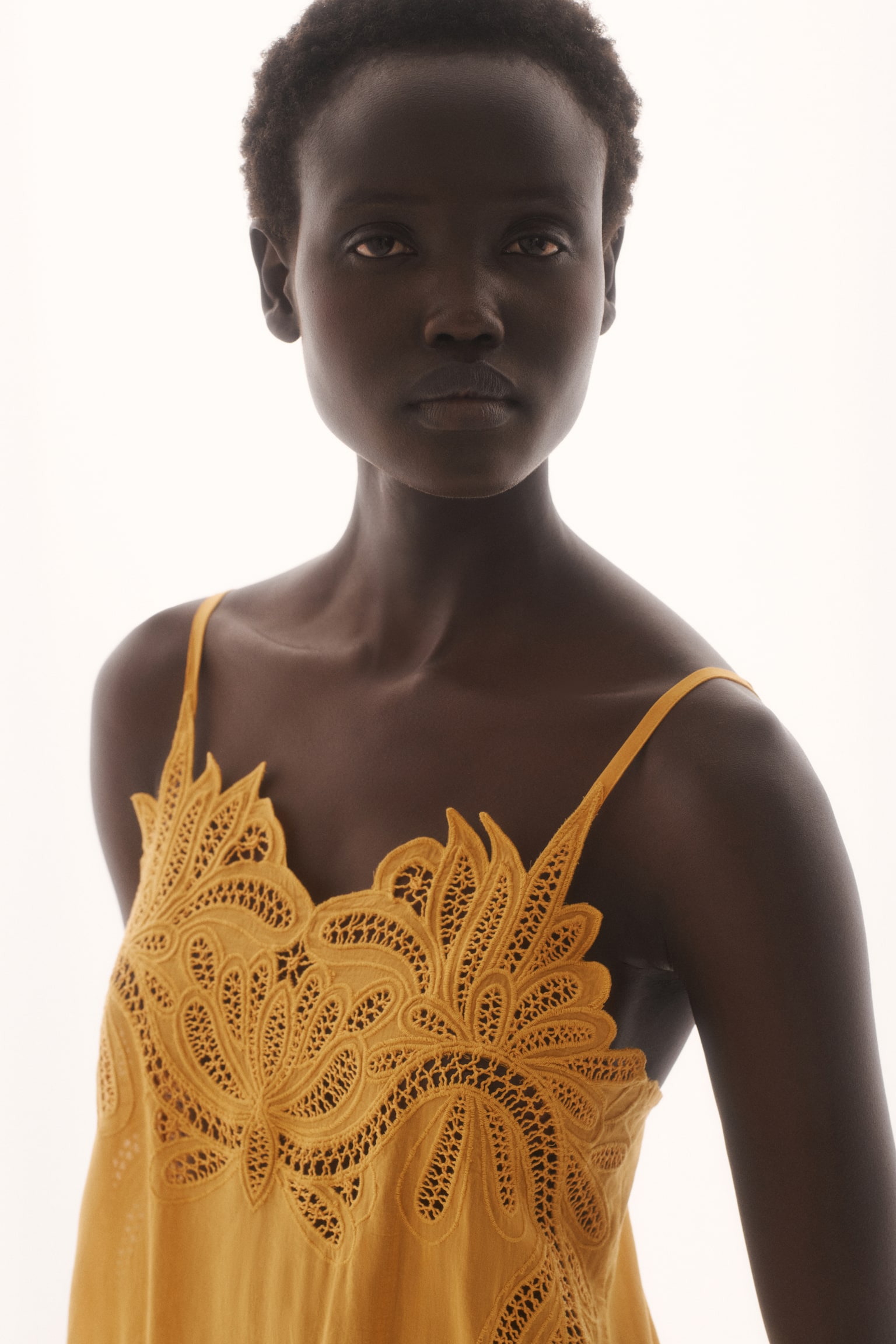 A short golden yellow dress is facing forward, featuring intricate broderie anglaise detailing across its chest and narrow, adjustable shoulder straps. A gathered seam is defining the waist, and a gently flared skirt is extending below. - 1