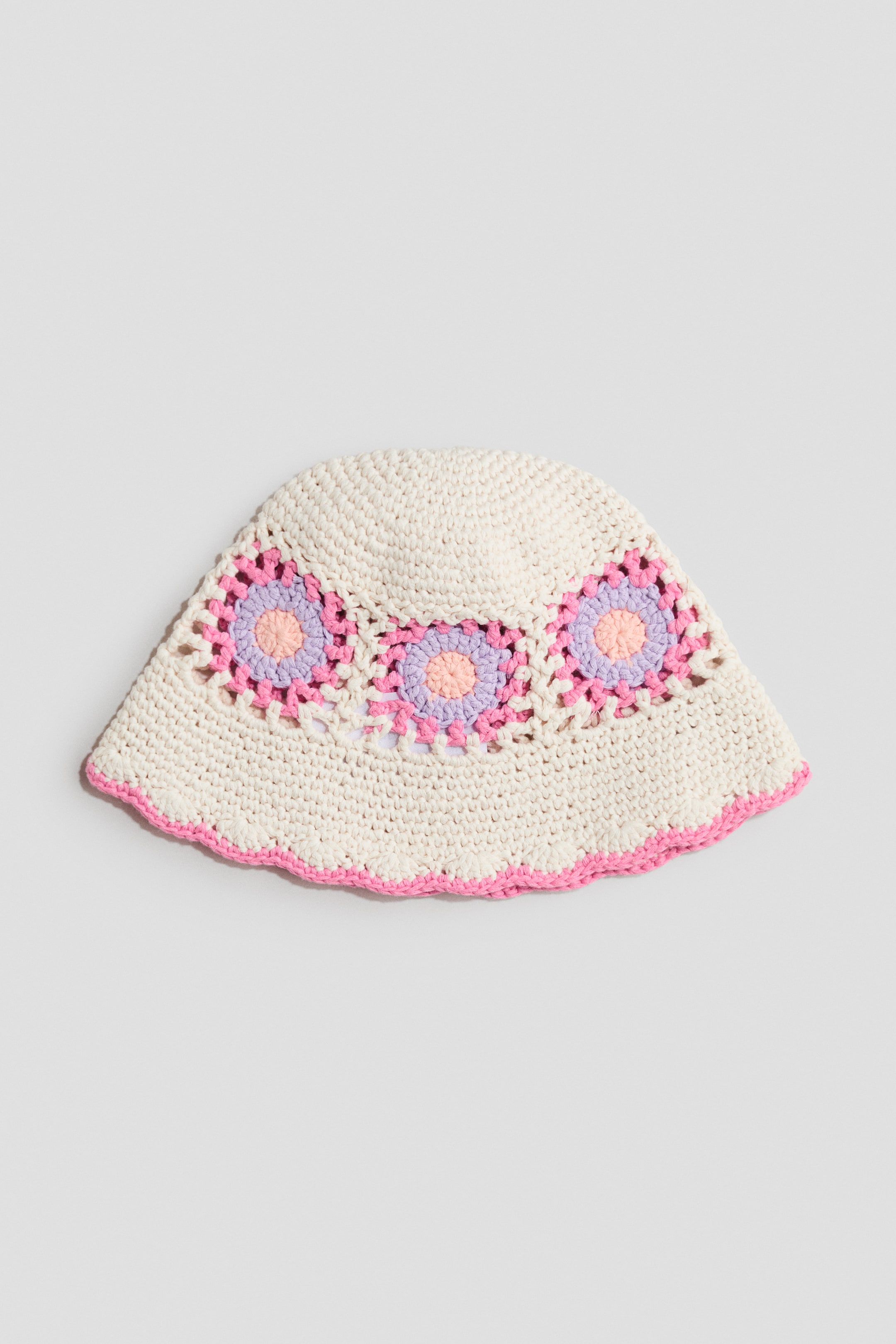 View larger image: A bucket hat is laid out flat, displaying a cream crochet-look knit cotton. It features a vibrant hole-knit pattern with three floral motifs in pink, purple, and peach around the crown, and a pink scalloped edge along the brim.