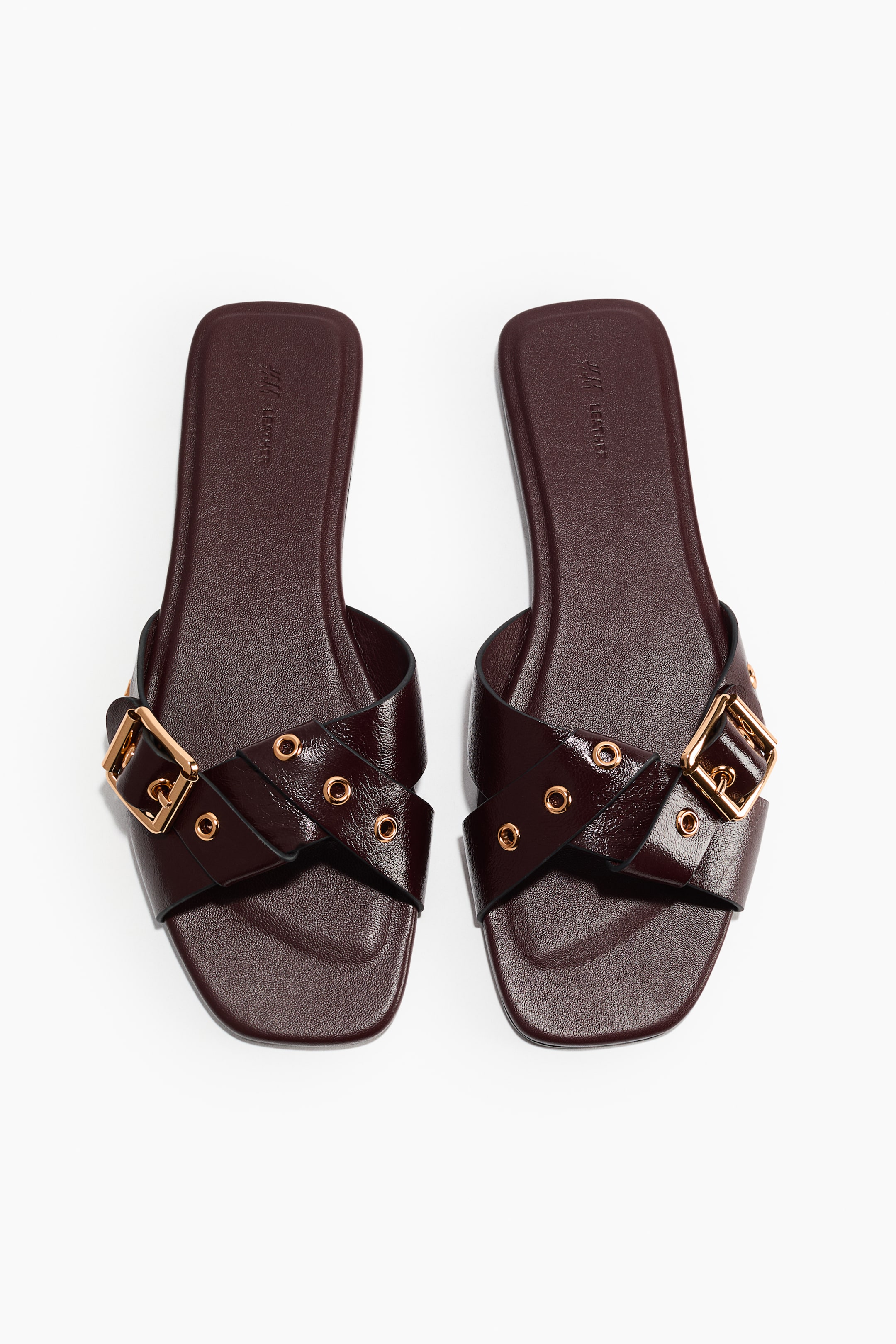 View larger image: A pair of burgundy flat sandals is facing front, featuring crisscrossing straps adorned with gold-toned eyelets. Each sandal is also displaying a gold-toned rectangular buckle on its outer side and showcasing a square toe with a flat sole.
