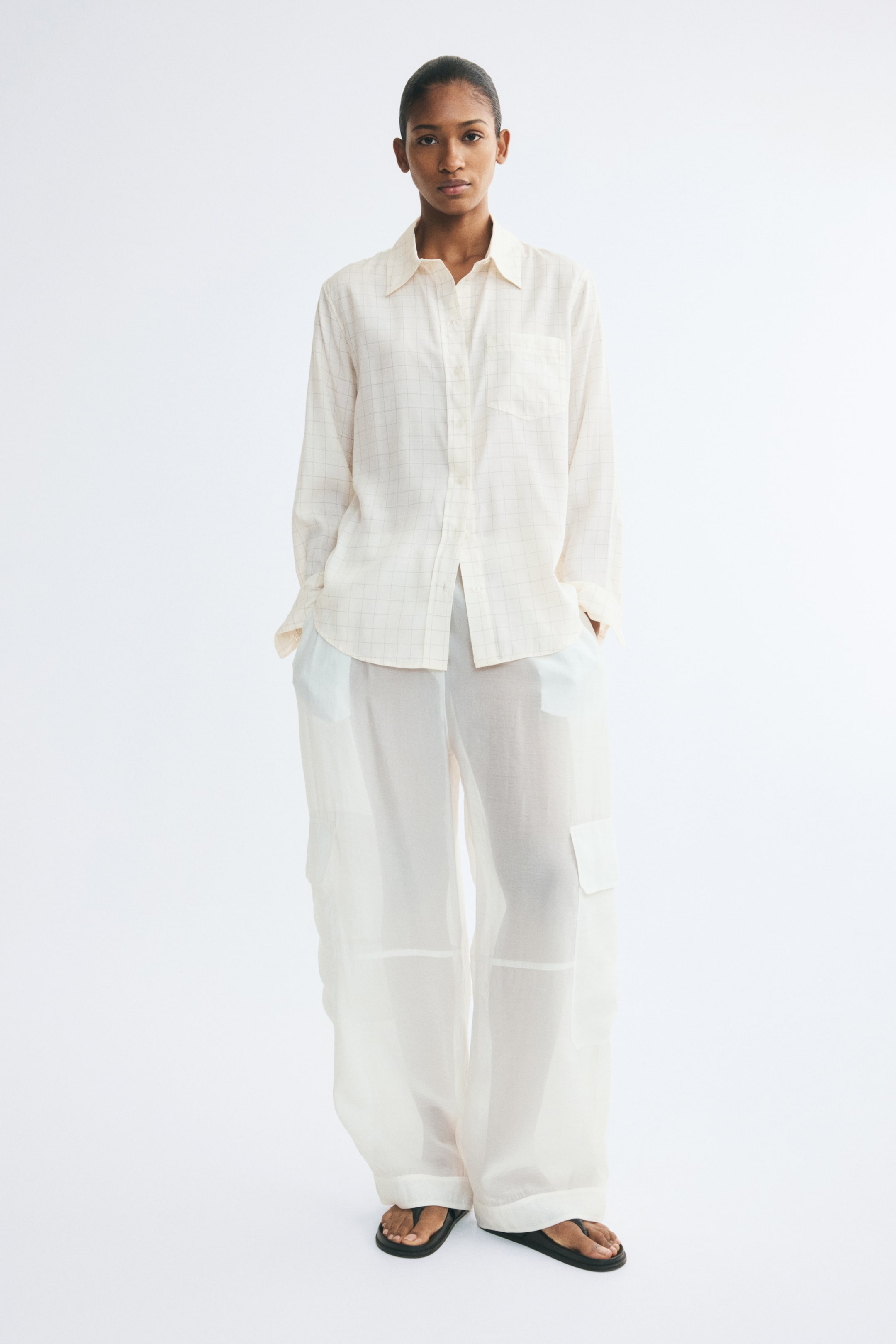 View larger image: A cream-coloured shirt with a grid pattern, featuring a collar, button-down front, and a single chest patch pocket, is worn by the model. The long-sleeved shirt is styled untucked over sheer, wide-leg cargo trousers, with the model standing with hands in the trouser pockets.