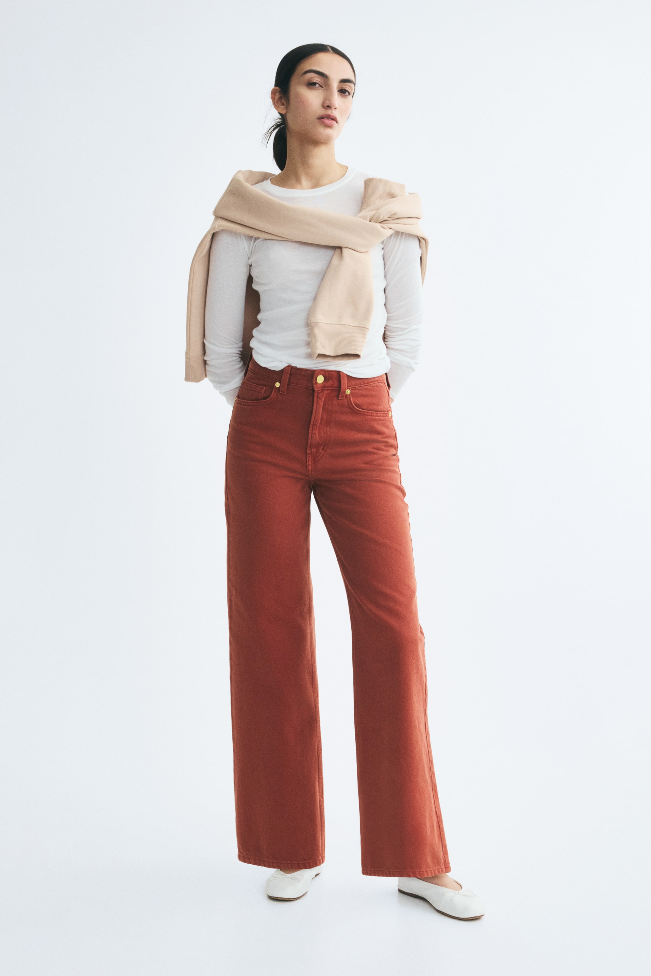 View larger image: These rust-red, high-waisted, straight-leg denim jeans are worn by the model and styled with a tucked-in white long-sleeve top and a beige sweater draped over the shoulders. The jeans fall straight to the ankle, resting just above the model’s white flat shoes.