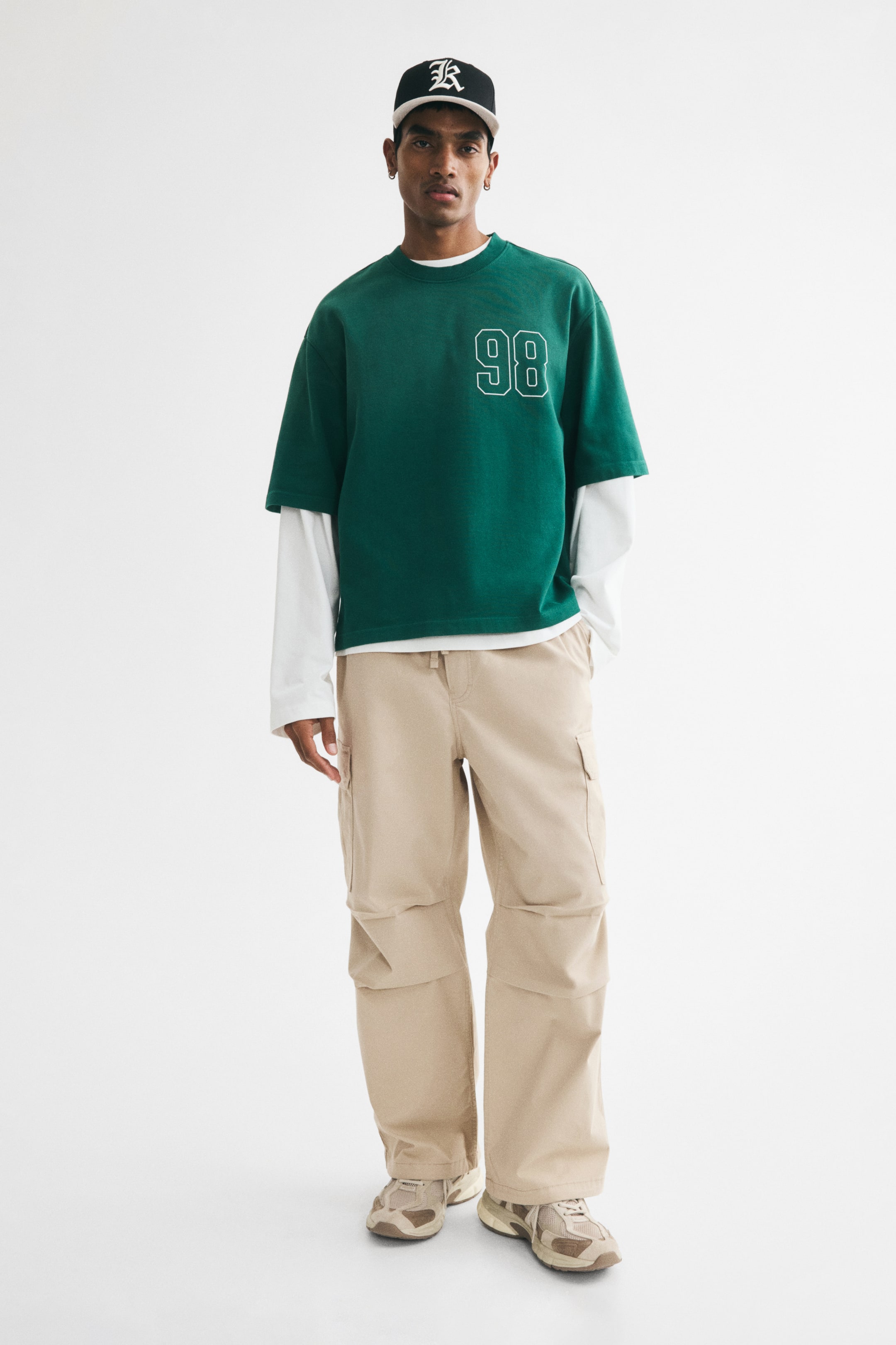 View larger image: The model is sporting cargo trousers crafted from cotton twill, showcasing a drawstring at the waist, a faux fly, diagonal side pockets, and patch leg pockets with a flap. These trousers are designed with knee pleats and offer a relaxed fit, with the model casually placing one hand in a side pocket.