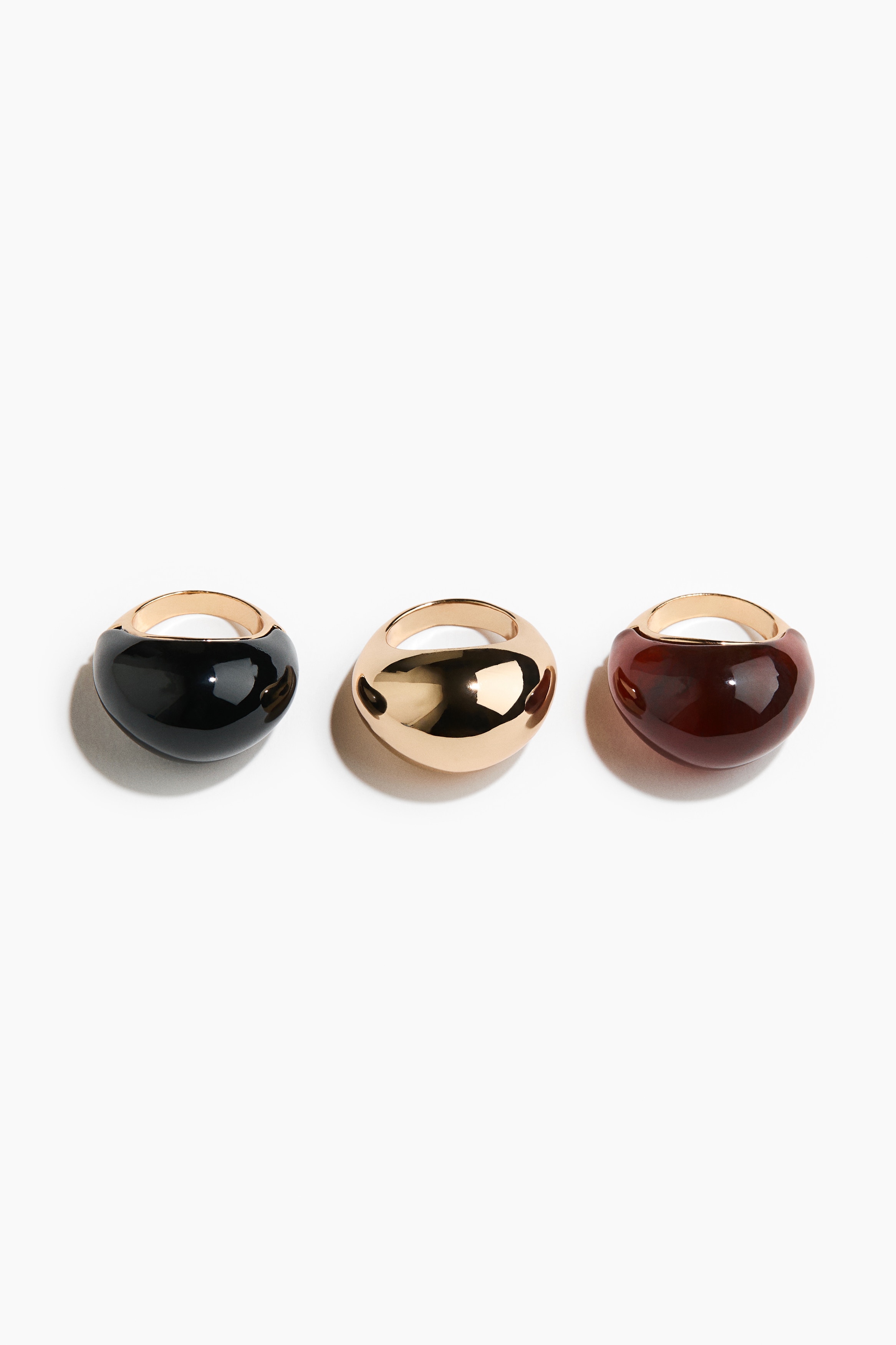 Chunky rings in metallic gold, glossy black, and translucent amber finishes are worn on the modelâs fingers, with their hands clasped together near the chest. The rings are styled with a dark brown, short-sleeved top.