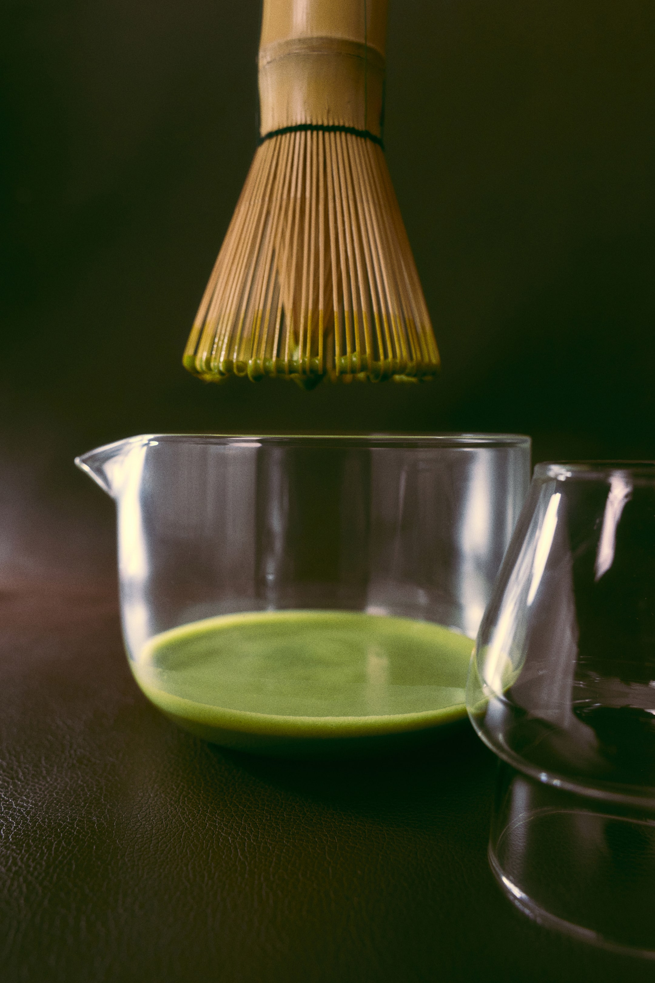 Ver imagen más grande: Se muestra un set de matcha con un batidor de bambú suspendido sobre un cuenco transparente de vidrio de borosilicato que contiene té verde batido. En primer plano, junto al cuenco con pico vertedor, descansa un soporte a juego de vidrio transparente para el batidor.
