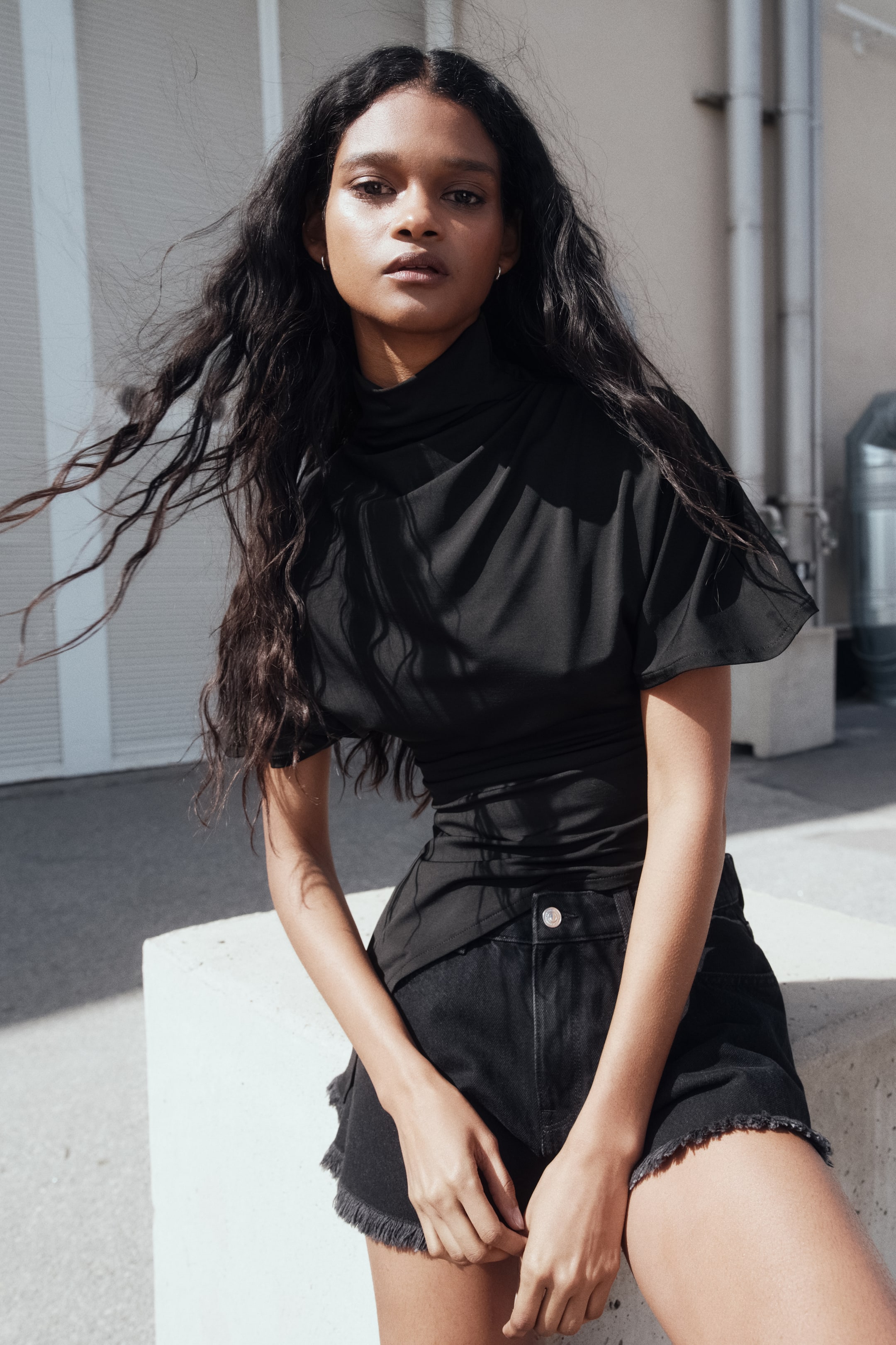 Ver imagem maior: Um top preto apresenta um decote drapeado em cowl e mangas curtas dolman, com a cintura ajustada inserida em calções de ganga preta. Os calções de ganga preta apresentam um corte de cintura alta e bainhas desfiadas.