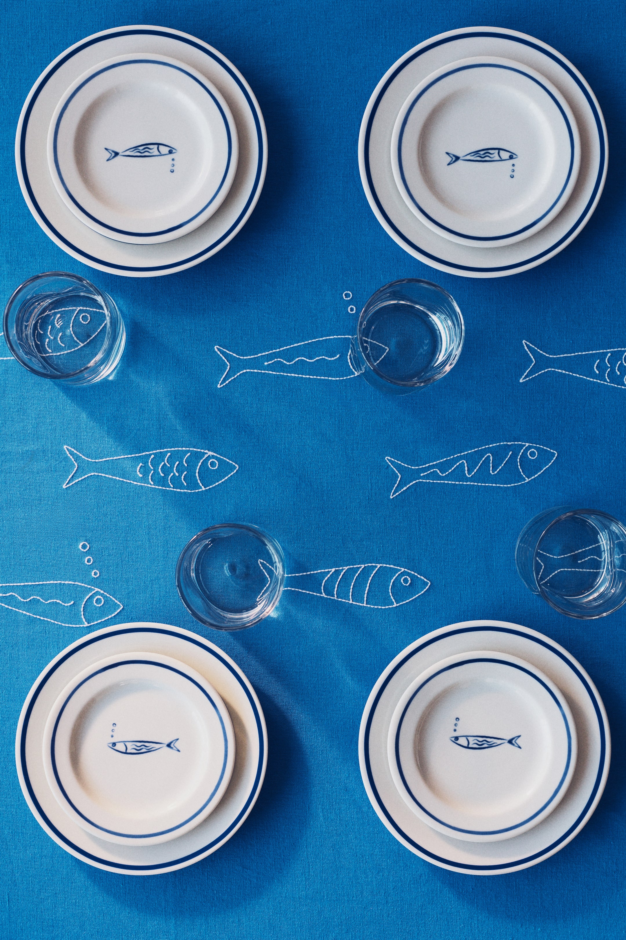 Grotere afbeelding bekijken: Vier serviesplaatsinstellingen worden getoond, elk bestaande uit een groot wit bord met een blauwe rand, gelaagd met een kleiner wit bord dat een blauw visontwerp en drie kleine blauwe cirkels weergeeft. Heldere drinkglazen zijn naast elke instelling gepositioneerd, allemaal rustend op een blauw tafelkleed met witte geborduurde visomtrekken.