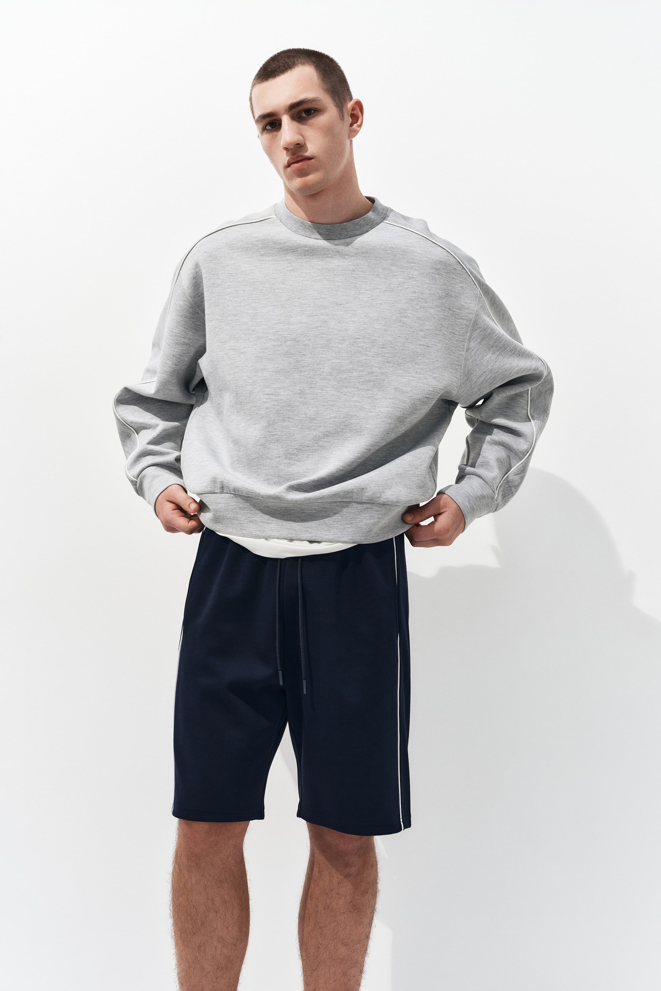 Grotere afbeelding bekijken: Een lichtgrijze sweatshirt met ronde hals heeft witte biezen langs de raglanmouwen, geribbelde boorden en een ontspannen pasvorm, gelaagd over een wit t-shirt. Marineblauwe shorts hebben een witte zijstreep en een tailleband met trekkoord.