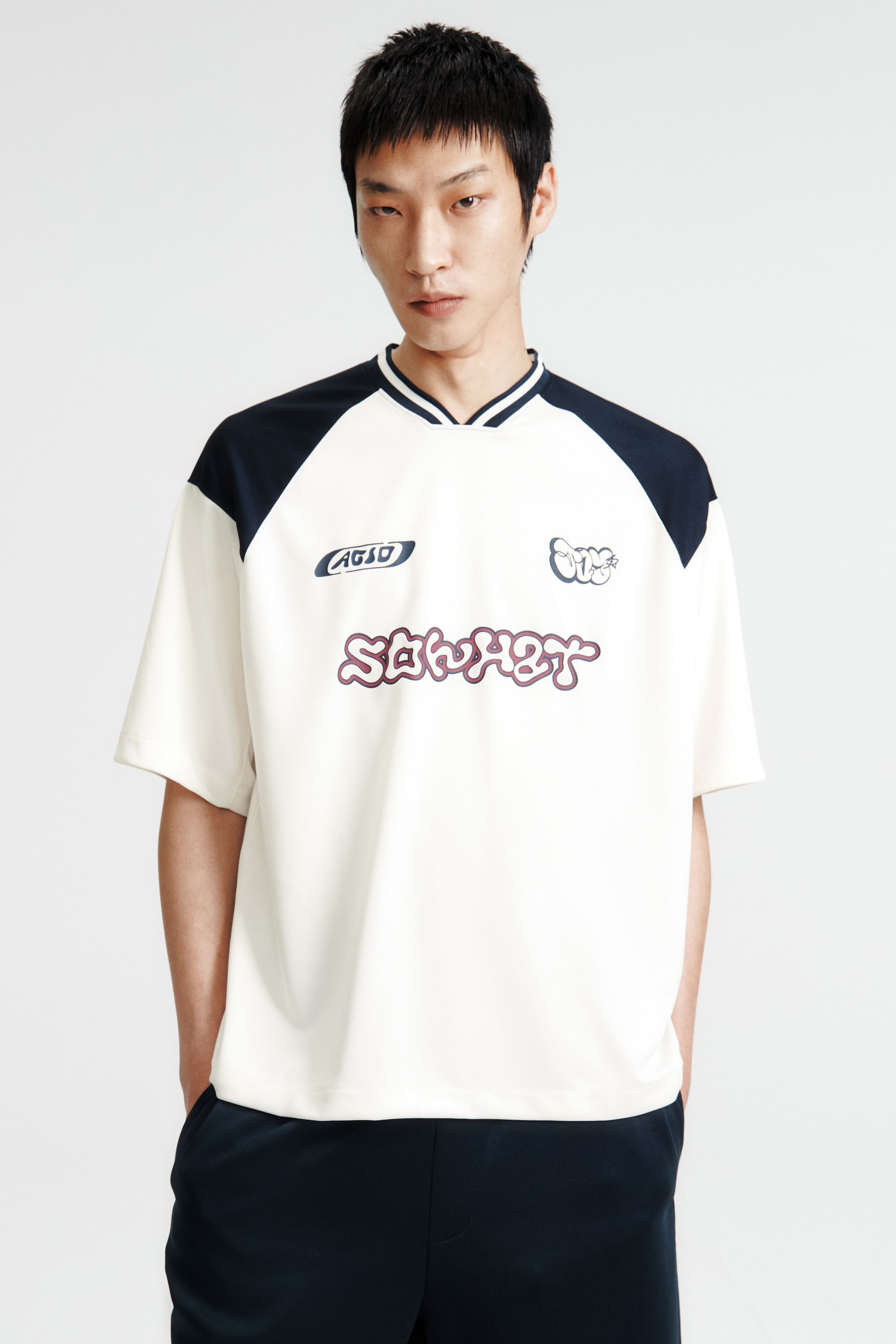 拡大画像を表示: モデルが着用しているTシャツは、クリーム色のメインボディにネイビーのショルダーヨーク、白いストライプが入ったリブ付きVネックが特徴です。前面には様々なプリントモチーフが施されており、半袖でストレートカットの裾があり、ゆったりとしたフィット感を演出しています。