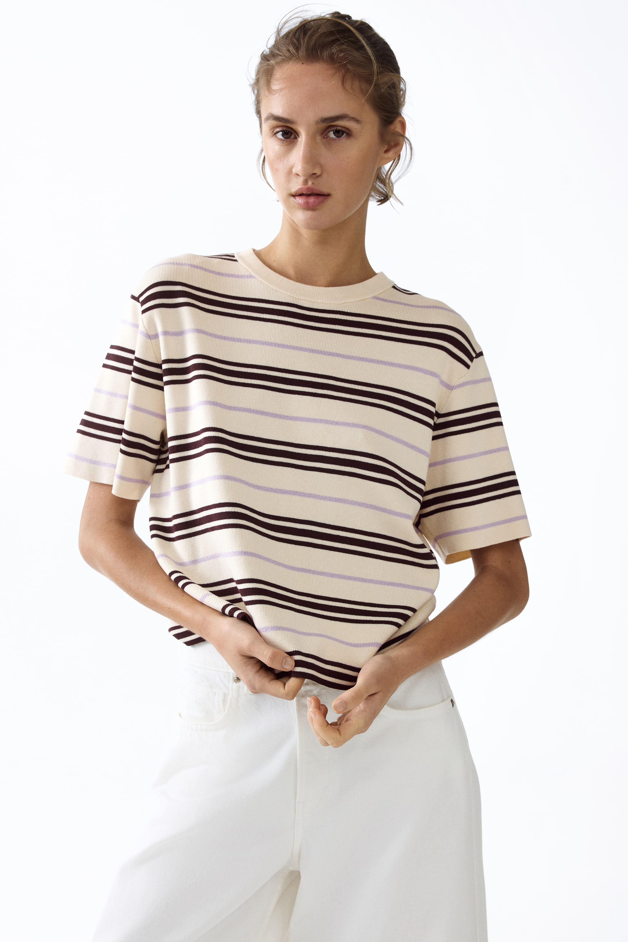View larger image: A cream-coloured t-shirt featuring horizontal stripes in dark brown and light purple, with a crew neck and short sleeves. The t-shirt is being slightly pulled at the hem, showcasing its relaxed fit and ribbed texture.