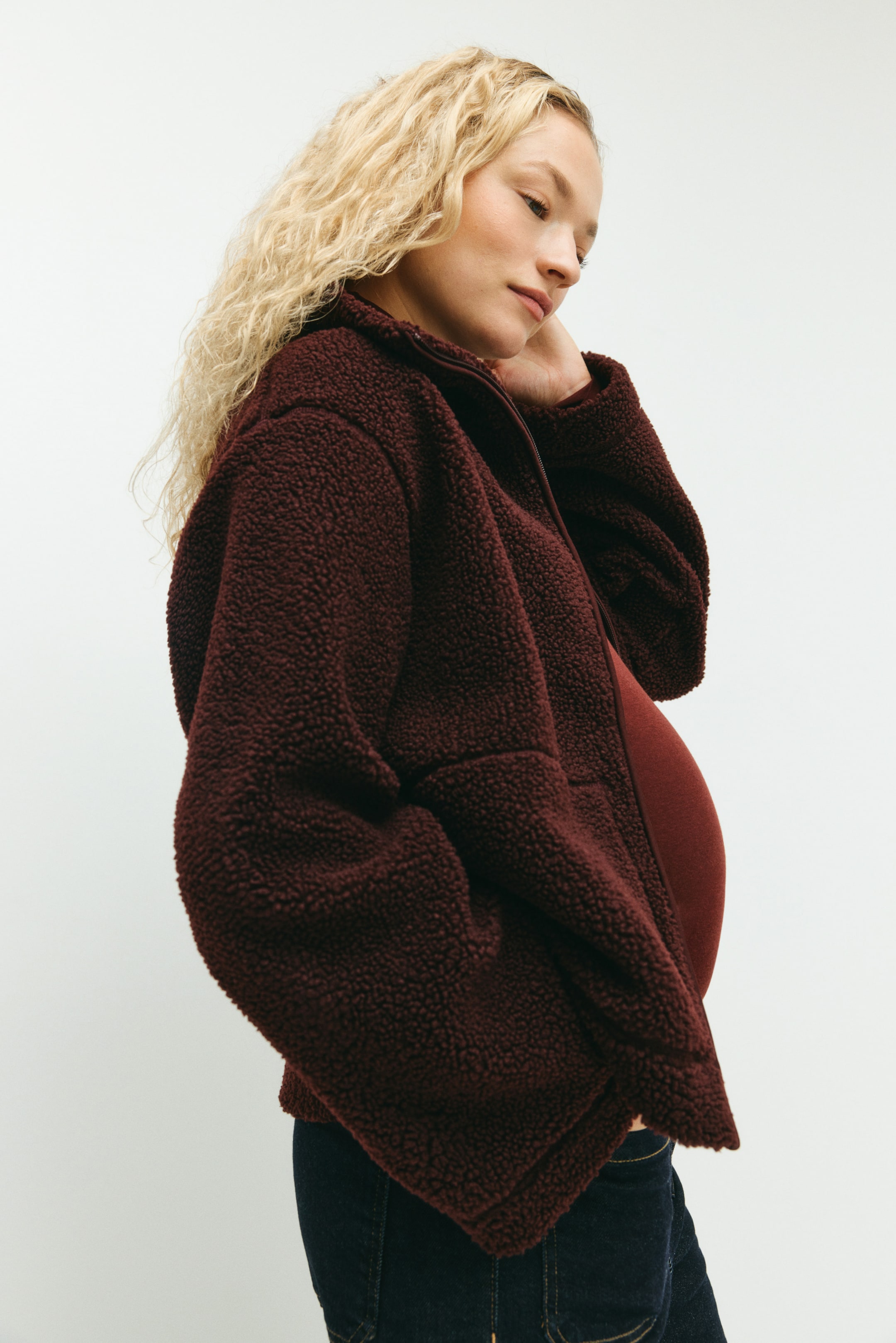 Se større billede: En mørkerød teddy-jakke bæres af modellen, hvor dens høje krave står op. Jakken's forlynlås er delvist åben, hvilket afslører en mørkerød top, og dens skuldre er synligt nedfaldne.