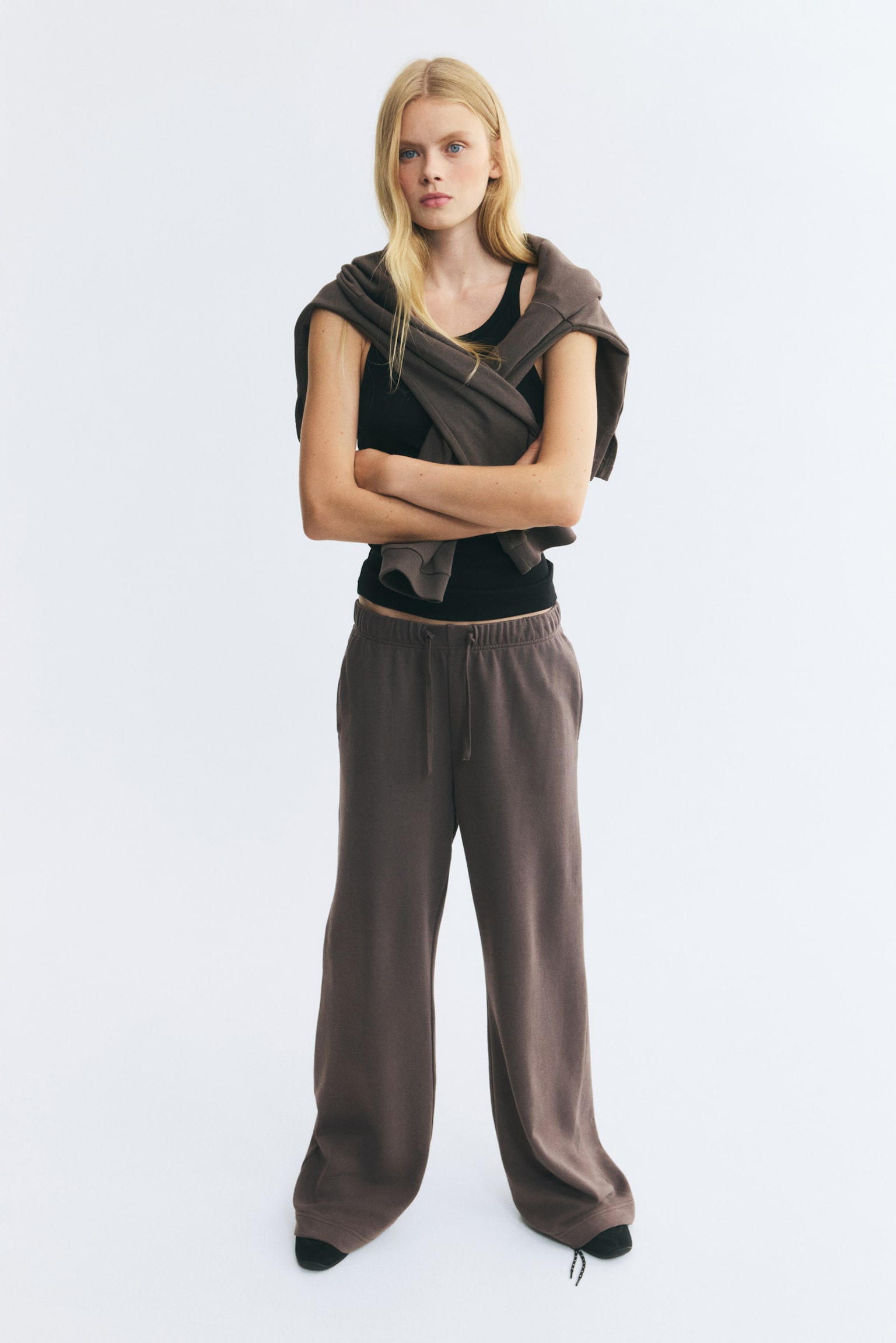 Brown track pants in a lightweight sweat fabric are being worn by the model, featuring an elasticated waistband with a drawstring and straight legs. The pants are styled with a black tank top and a matching brown sweatshirt draped over the shoulders. - 1