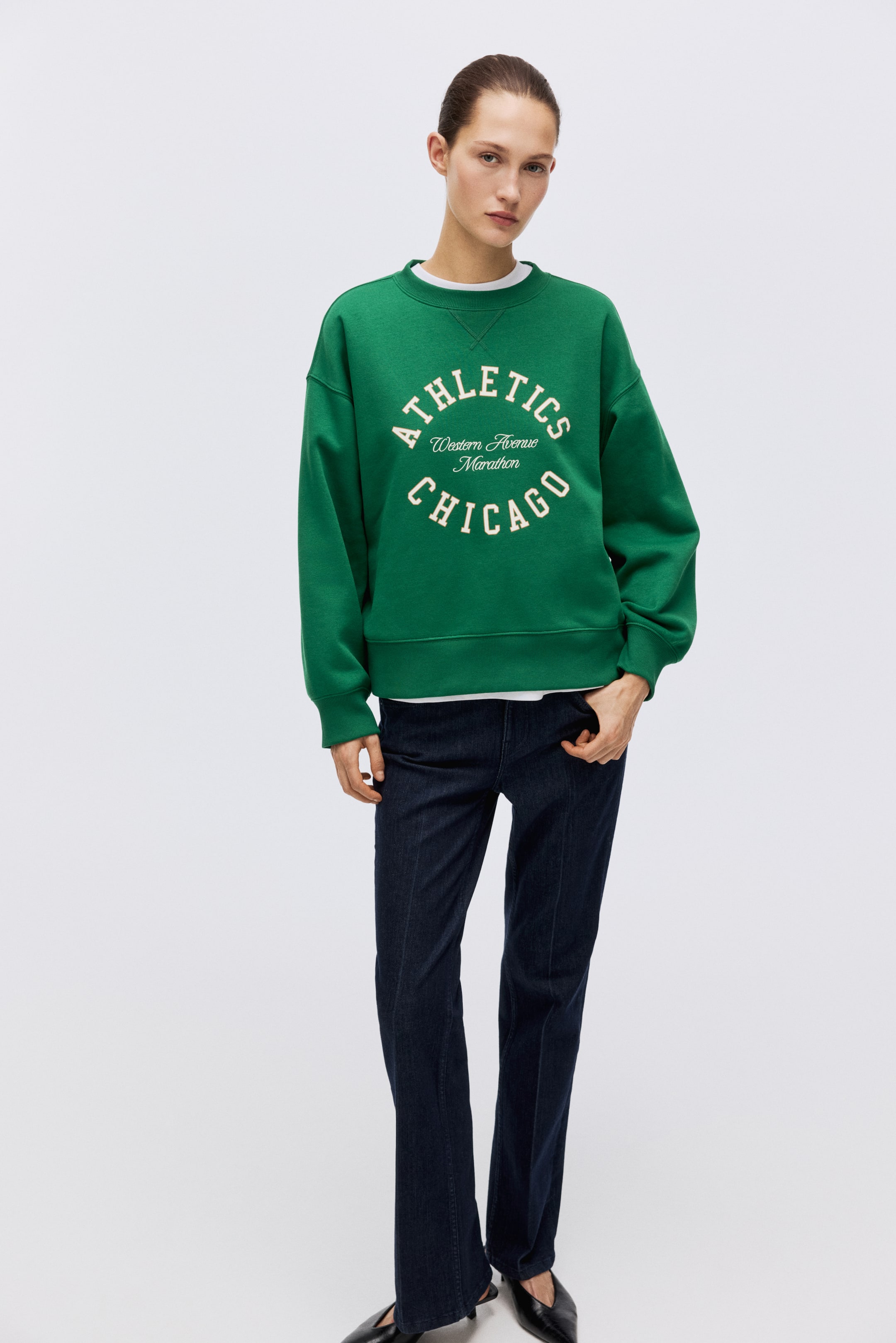 View larger image: A green crew-neck sweatshirt displays white arched text 'ATHLETICS' above 'Western Avenue Marathon' and 'CHICAGO' on the front. It features ribbed cuffs and a ribbed hem, with a white T-shirt layered underneath at the neckline, and is angled slightly to the left to showcase its relaxed fit.