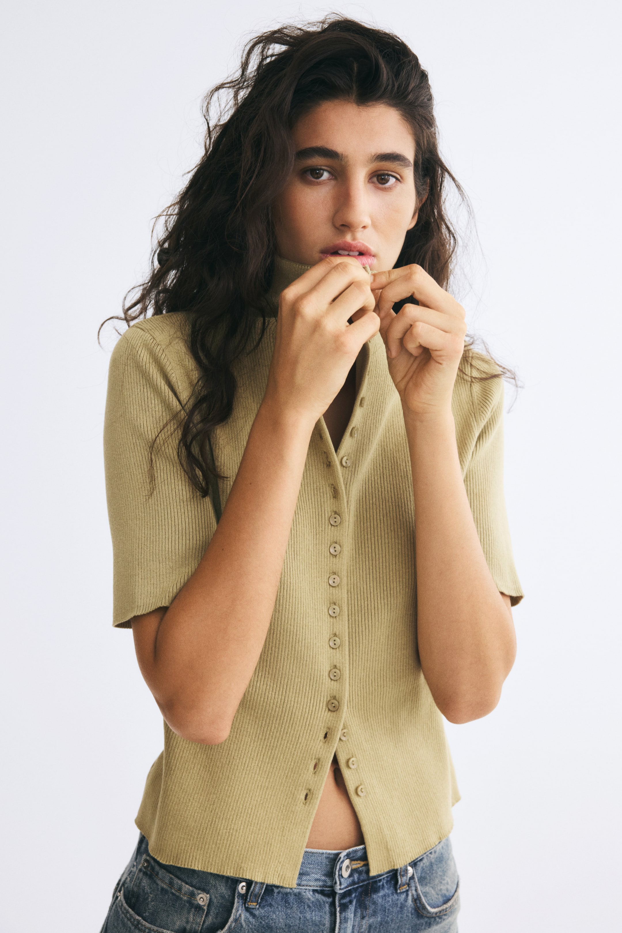 View larger image: A light green, short-sleeved ribbed-knit cardigan is being worn by the model, who is adjusting the high-neck button closure with both hands. The fitted cardigan is styled with blue denim jeans.