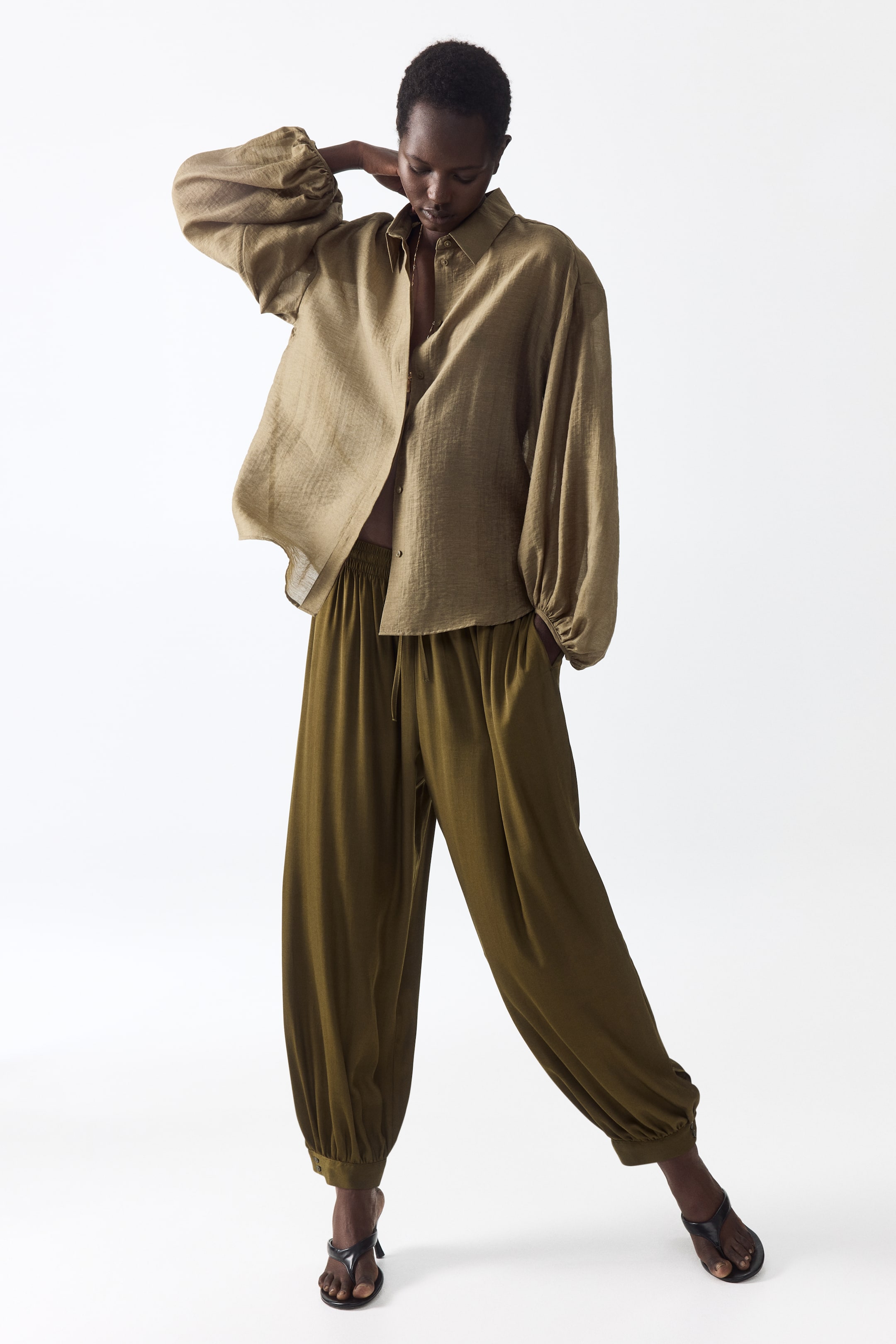 View larger image: The olive green matching set showcases a loose-fitting, crinkled-texture shirt, worn open, with voluminous sleeves gathered at the cuffs. The coordinating wide-leg trousers are styled with a visible drawstring waist and gathered ankles.
