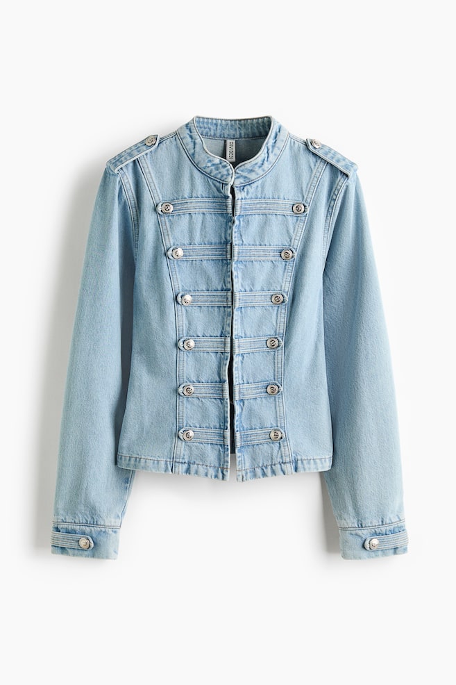 Eine hellblaue Jeansjacke ist frontal ausgerichtet und verfügt über einen Stehkragen und Epauletten, die jeweils einen silberfarbenen Knopf zeigen. Die Vorderseite präsentiert fünf horizontale Denim-Bänder, die jeweils mit zwei silberfarbenen Zierknöpfen auf beiden Seiten der mittigen Öffnung versehen sind, während die langen Ärmel in Manschetten mit einem Knopf enden. - 1