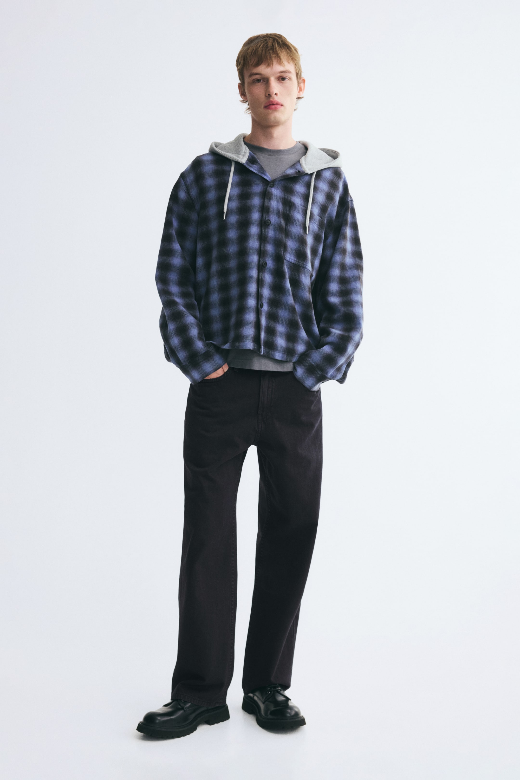 View larger image: Dark cotton twill 5-pocket pants in a relaxed fit with straight legs are worn by the model, who is standing with one hand in a front pocket. The pants are styled with a blue and black plaid hooded shirt and black chunky shoes.