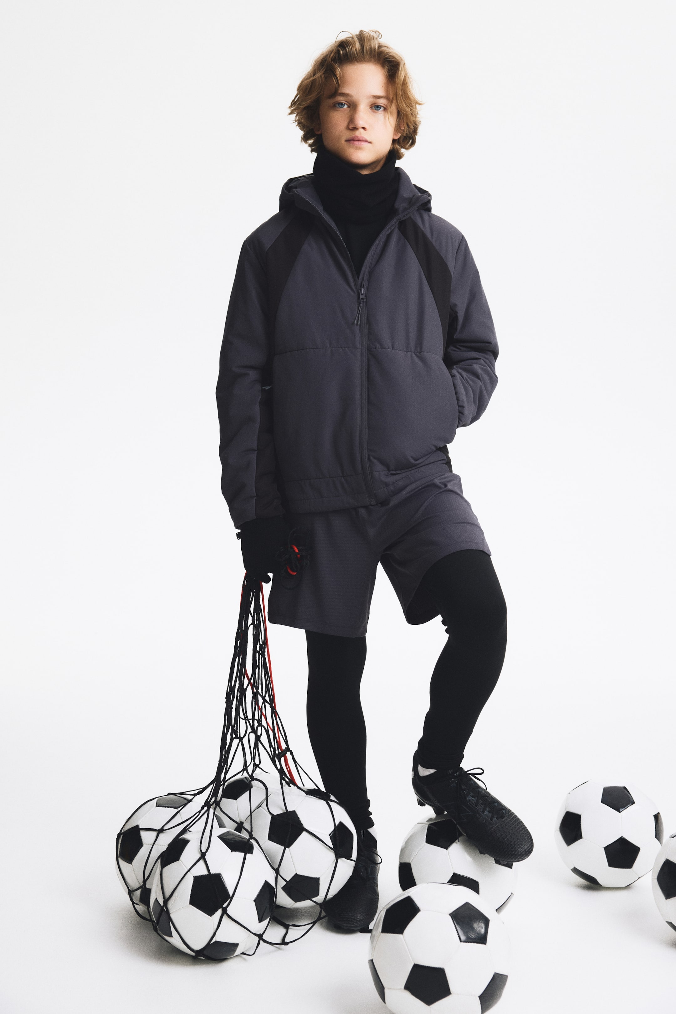 Ver imagen más grande: Un niño lleva un conjunto deportivo gris oscuro, de frente, formado por una chaqueta con capucha y cremallera, con piezas negras en los hombros, y unos pantalones cortos holgados a juego. La chaqueta se lleva sobre un top negro de cuello alto, mientras que los pantalones cortos se combinan con leggings negros de largo completo y se completan con zapatillas deportivas negras.