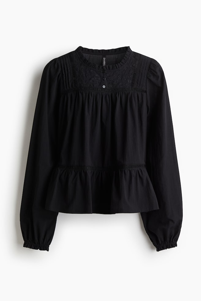 A black blouse is being presented FRONT, made from an airy cotton weave. It is featuring a frilled collar, buttons at the top, a yoke with pintucks and embroidery on the front, and ladder lace insets on the front. Long balloon sleeves with frilled, elasticated cuffs and a tier with a ladder lace inset at the front of the waist are also visible. - 1