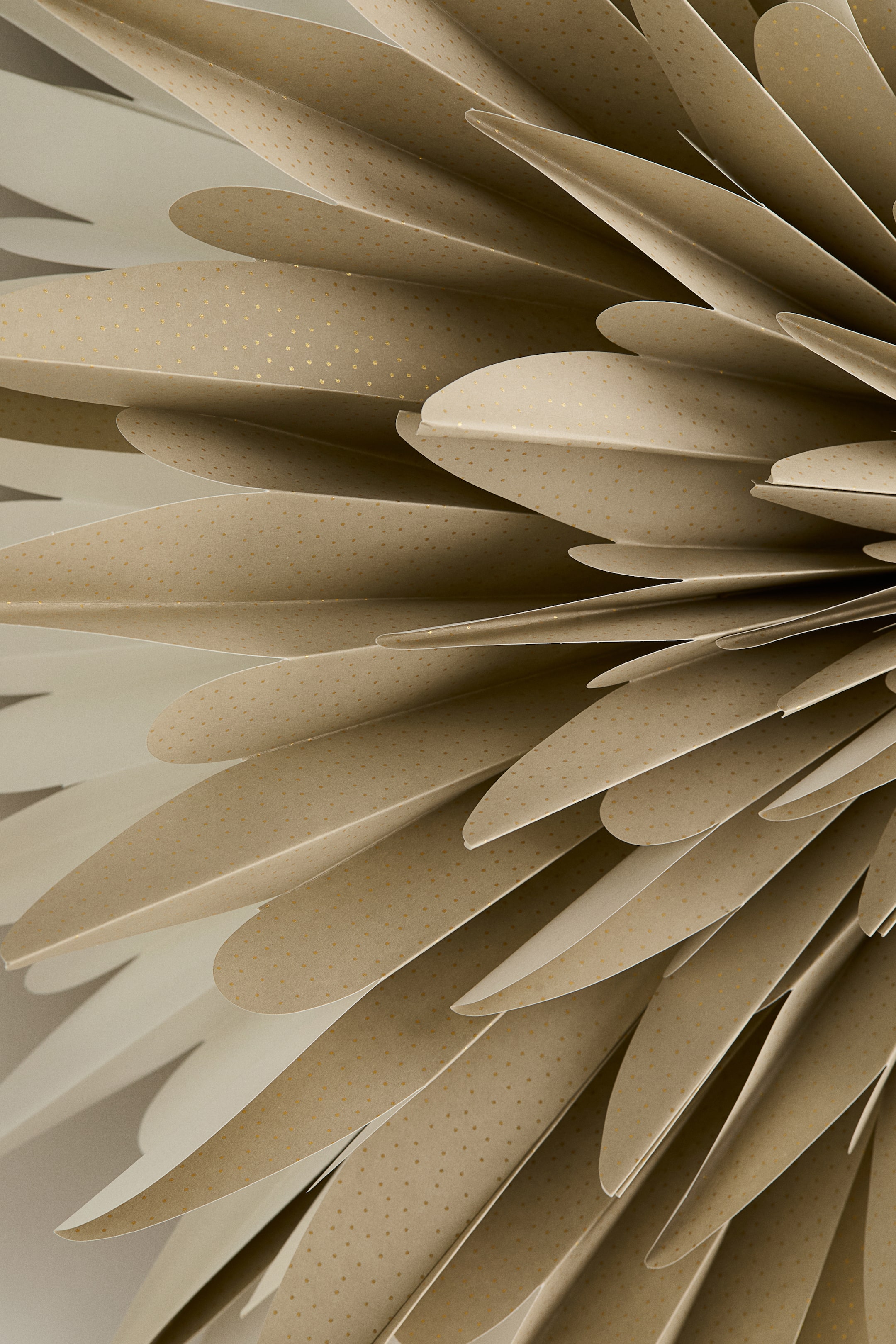 Se større billede: En papir stjerne præsenterer sit indviklede, lagdelte design, sammensat af adskillige plisserede segmenter med blødt afrundede kanter. Hvert segment har en lys beige farve med diskrete gyldne pletter, som tilsammen danner et fjeragtigt, radielt mønster.
