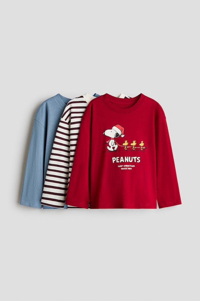 Three long-sleeve t-shirts are hanging in a layered, front-facing presentation, displaying a solid blue tee, a white and dark red striped tee, and a red tee. The red tee in the front is showcasing a round, rib-trimmed neckline and a printed graphic of Snoopy and Woodstock with the text "PEANUTS." - 1