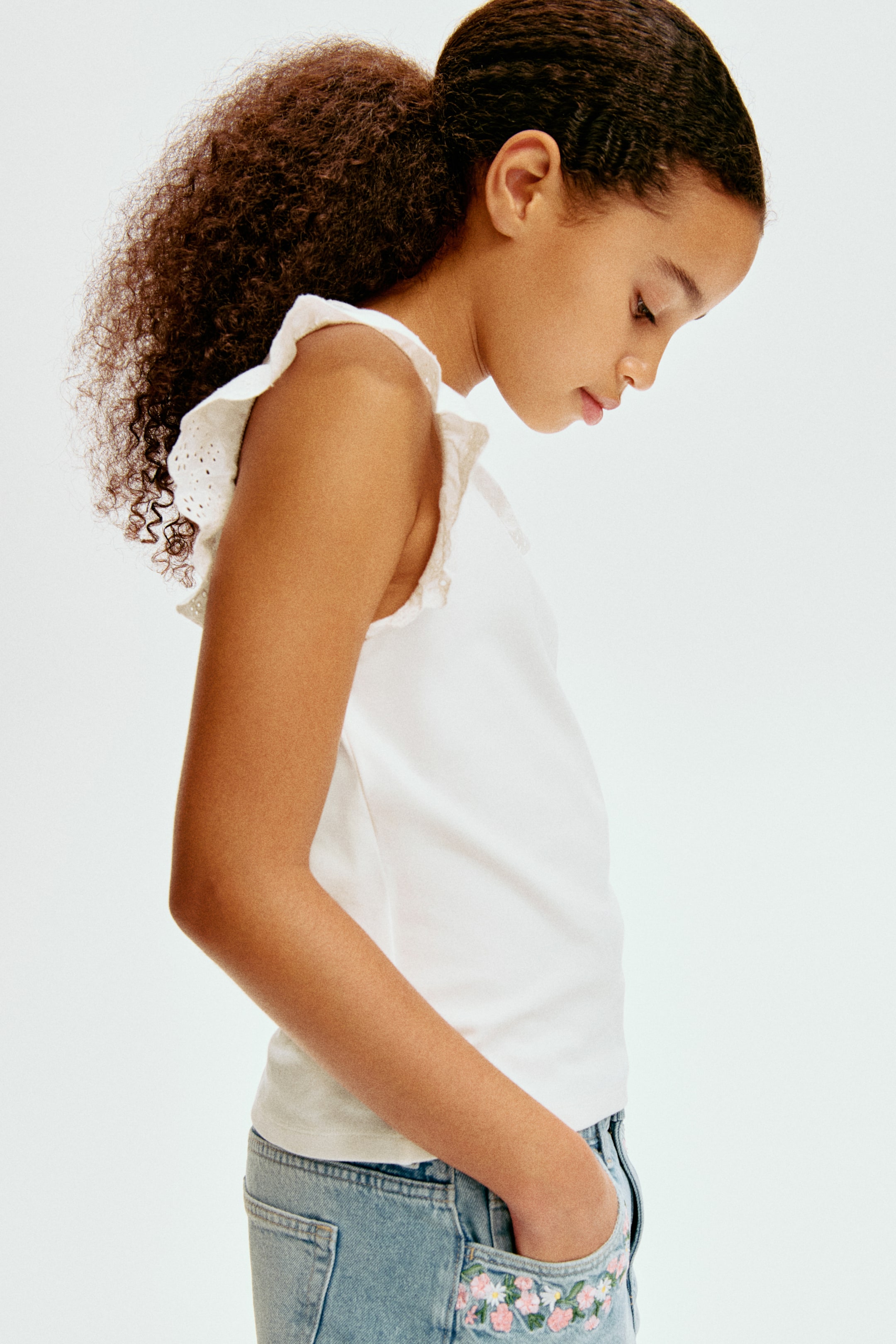 Grotere afbeelding bekijken: Een witte mouwloze top met ruches en geborduurde gaatjes bij de armsgaten wordt van opzij gedragen, gecombineerd met lichtblauwe denim shorts met een geborduurd bloemenmotief op de voorzak.