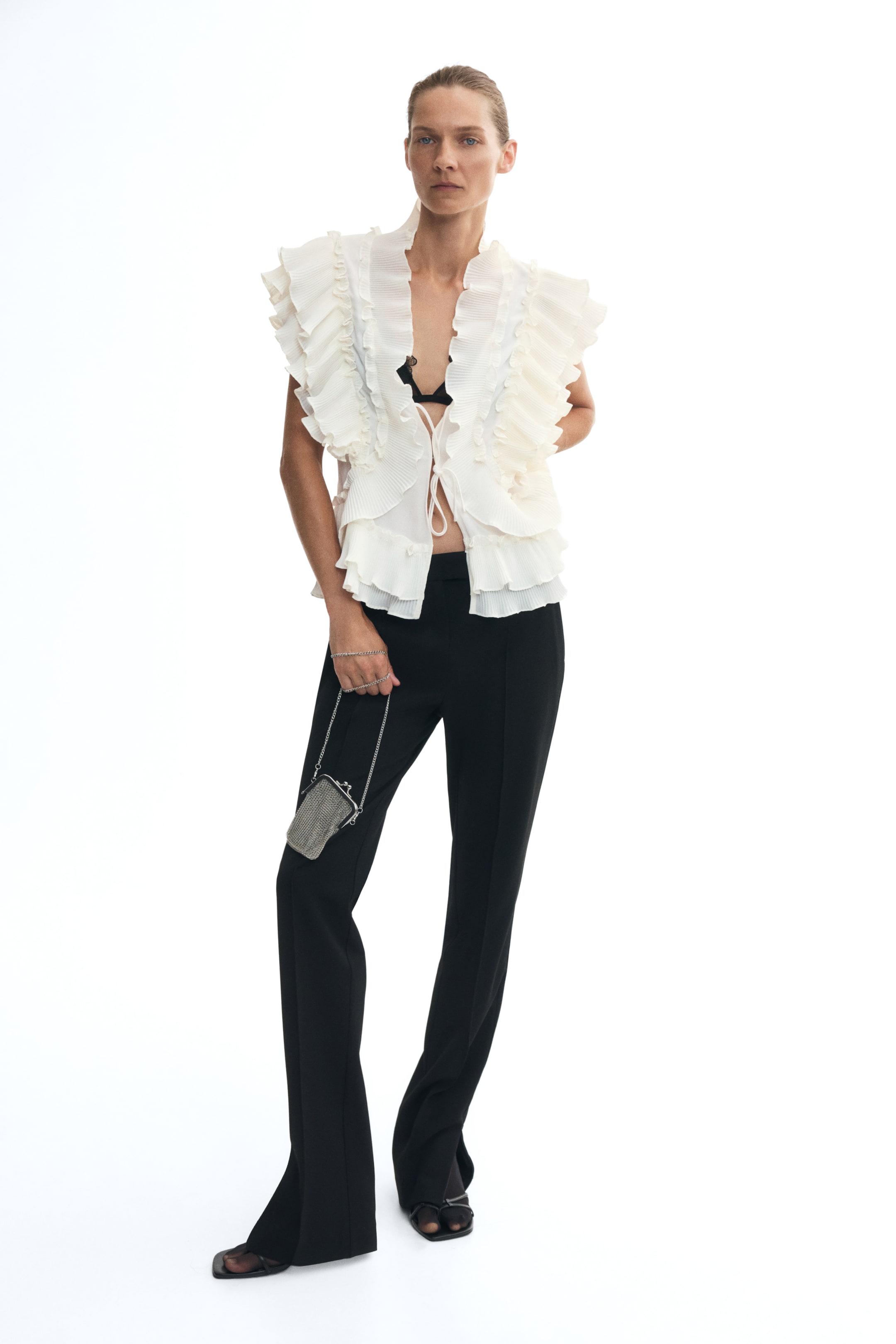 View larger image: A cream-colored sleeveless blouse featuring tiered flounces and a frill collar is worn by the model over a black bra. The open-front blouse is secured with narrow ties and paired with black trousers, while the model holds a small metallic chain handbag.