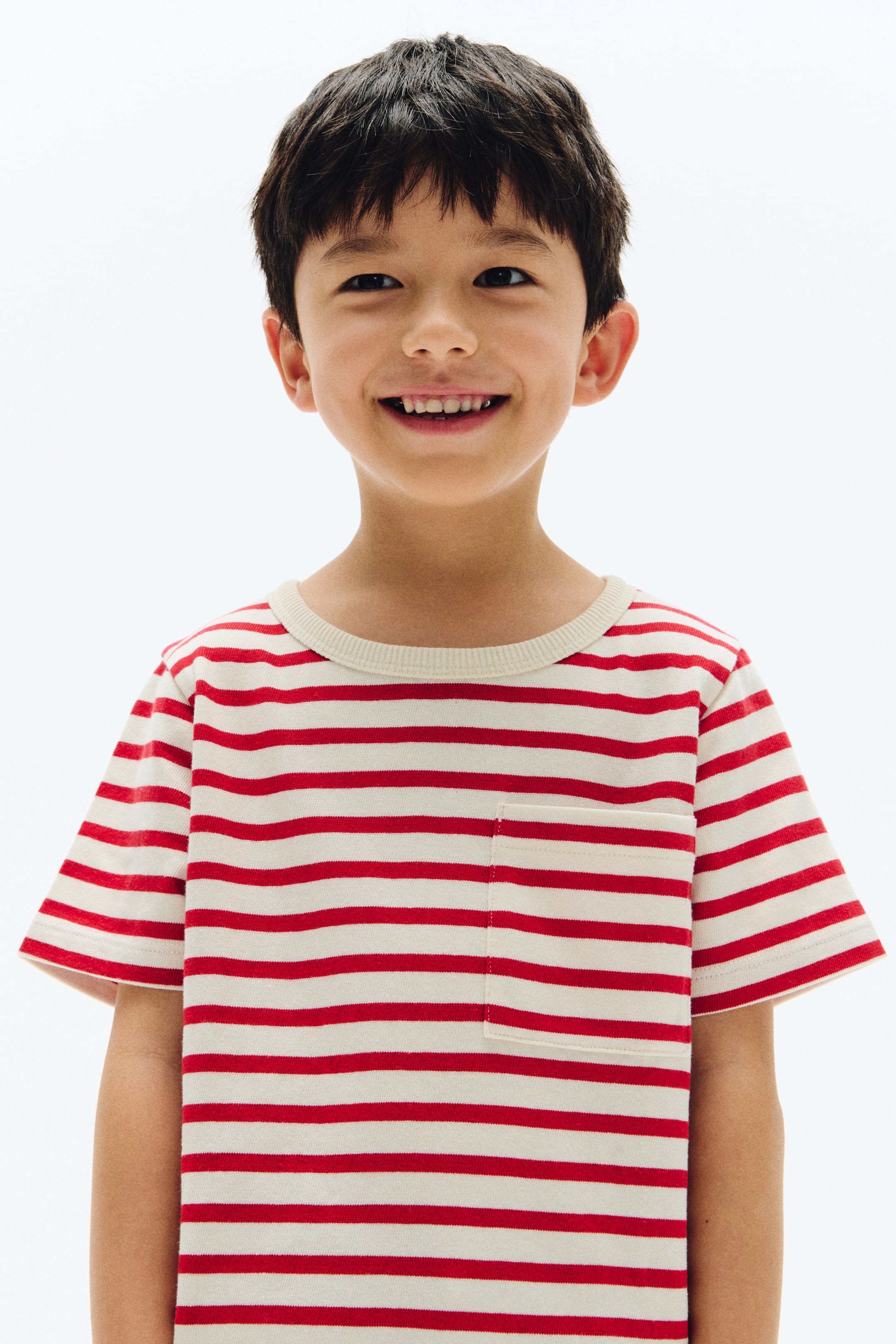 View larger image: A red and cream striped T-shirt is shown from the front, featuring a round ribbed neckline and short sleeves. It has horizontal red and cream stripes and a small patch pocket on the left chest, designed to fit loosely on a child.