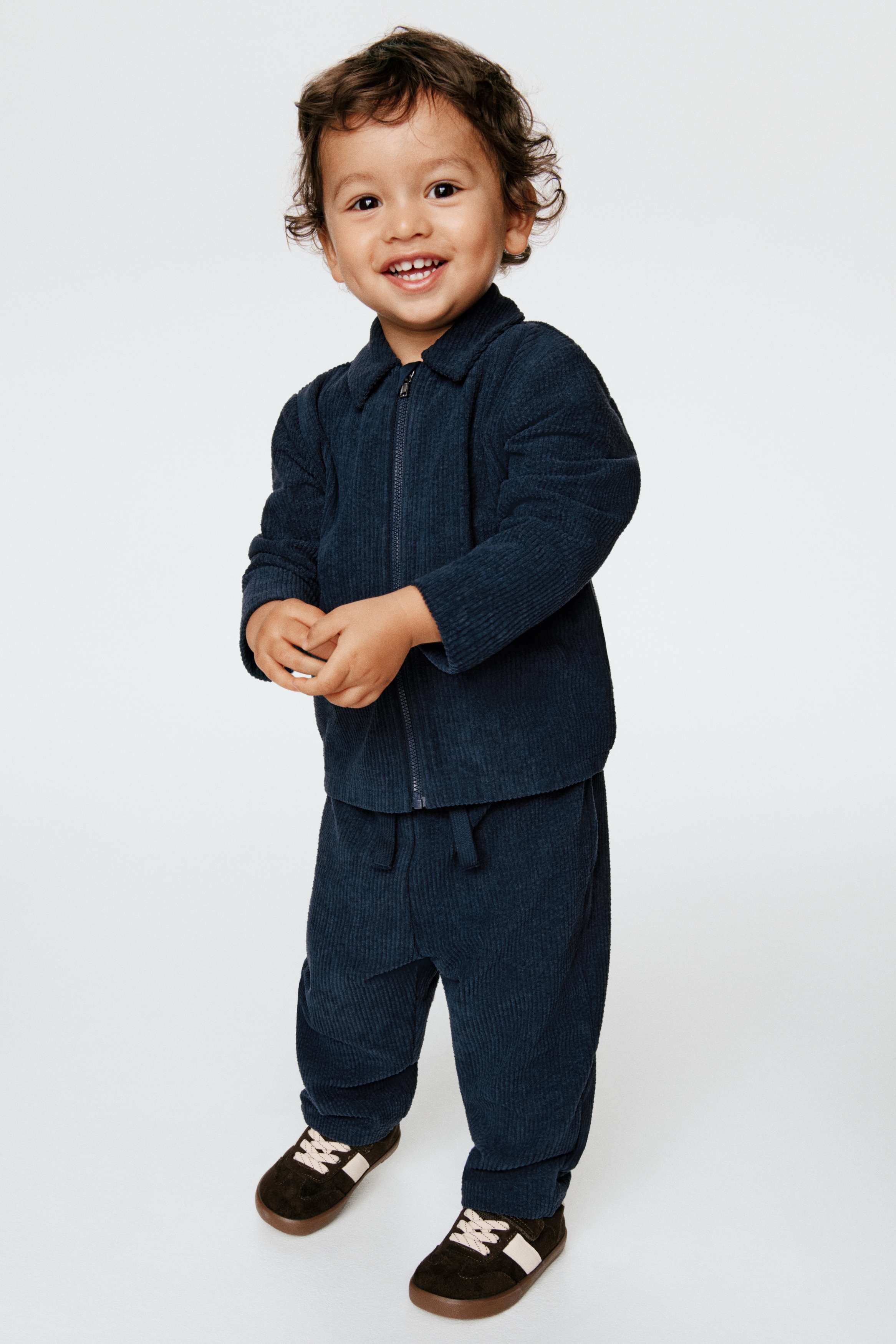A children's navy blue corduroy clothing set is displayed in a front view, featuring a collared, long-sleeved zip-up jacket and matching pull-on trousers. The jacket is left open, showing a plain white crewneck T-shirt layered underneath.