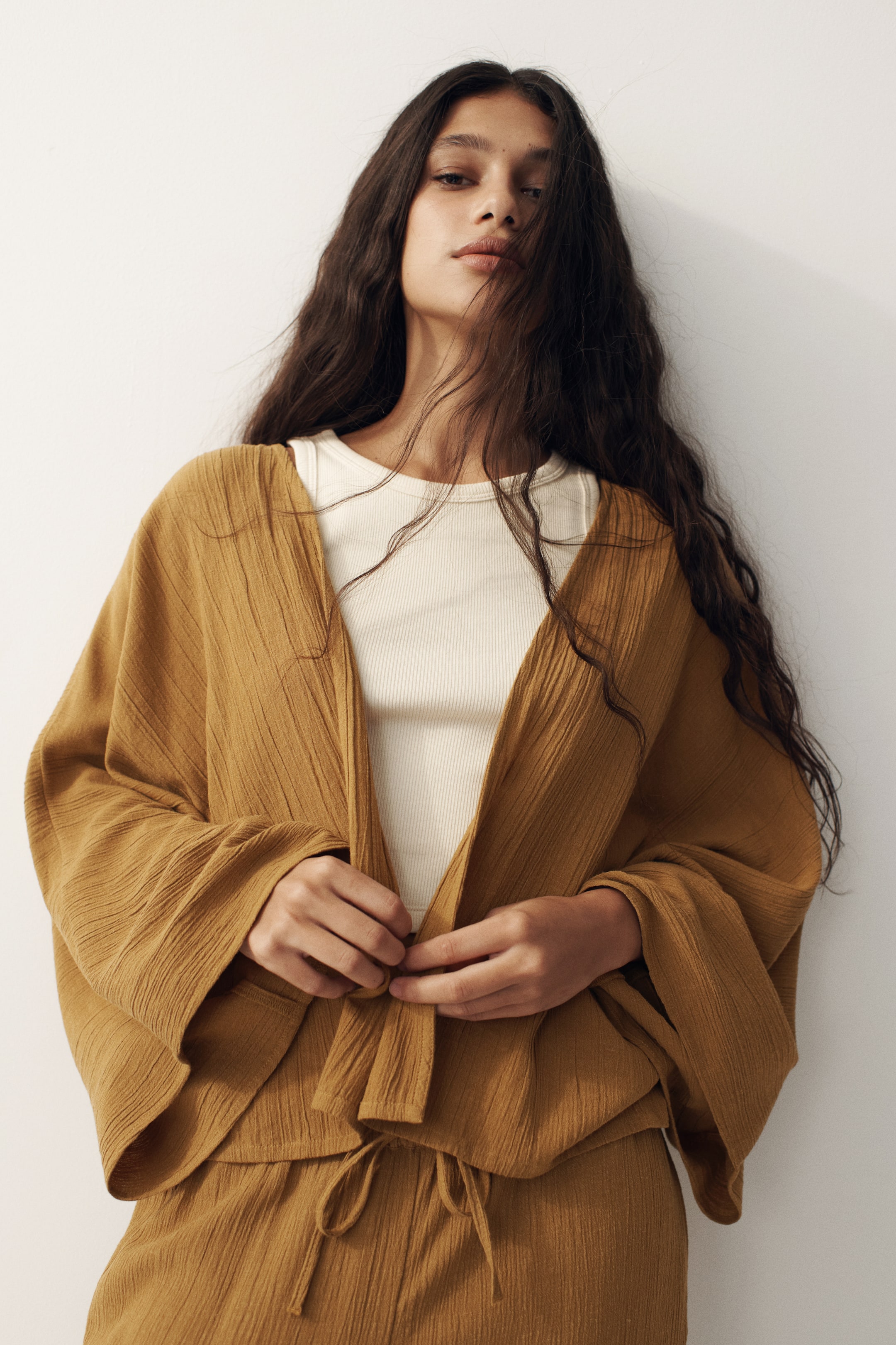 View larger image: A mustard-coloured textured open-front top with wide, flowing sleeves is being worn, revealing an off-white ribbed tank top underneath. The top is tied at the waist, and matching textured trousers with a drawstring waist are also visible.