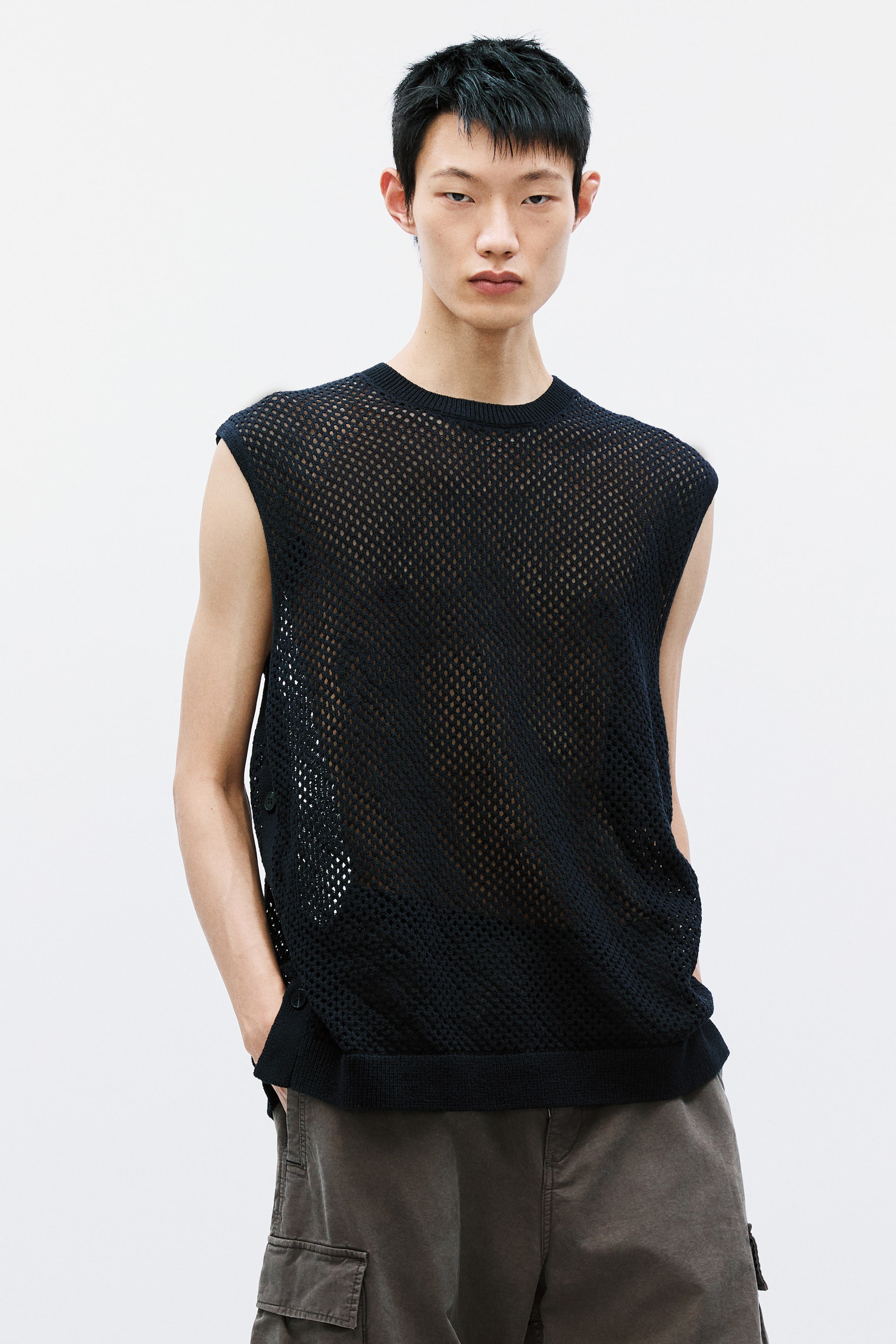 View larger image: Presenting a black mesh sleeveless top, showcasing a sheer, open-knit design with a ribbed crew neck and hem. It features side vents, each secured with two buttons, adding to its structural elegance.
