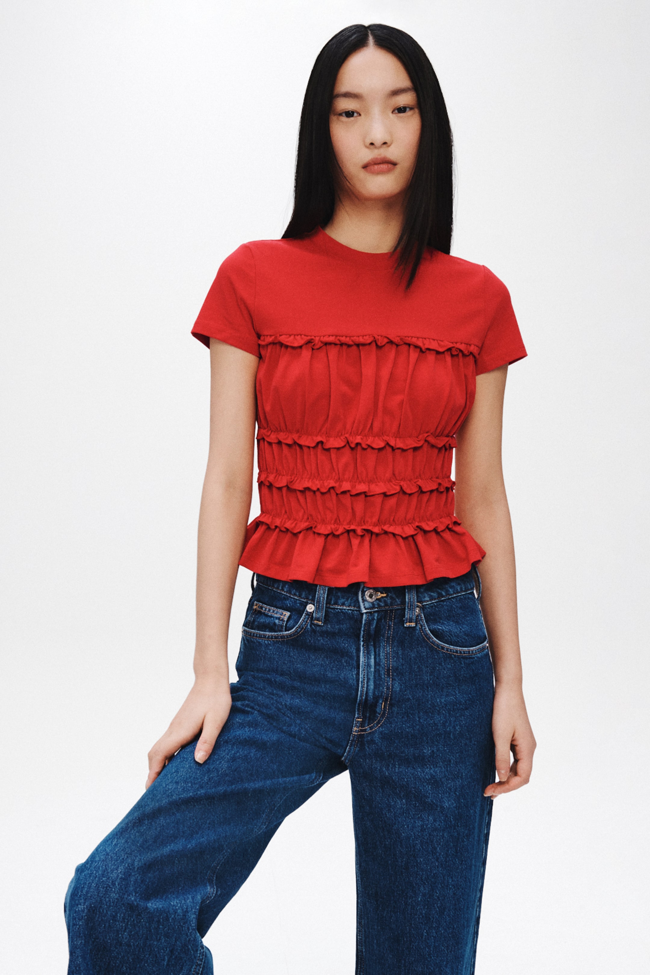 View larger image: The model is wearing a red fitted short-sleeved top with a round neckline, gathered tiers with frill trims at the front and a flared hem. The top is styled with blue jeans.