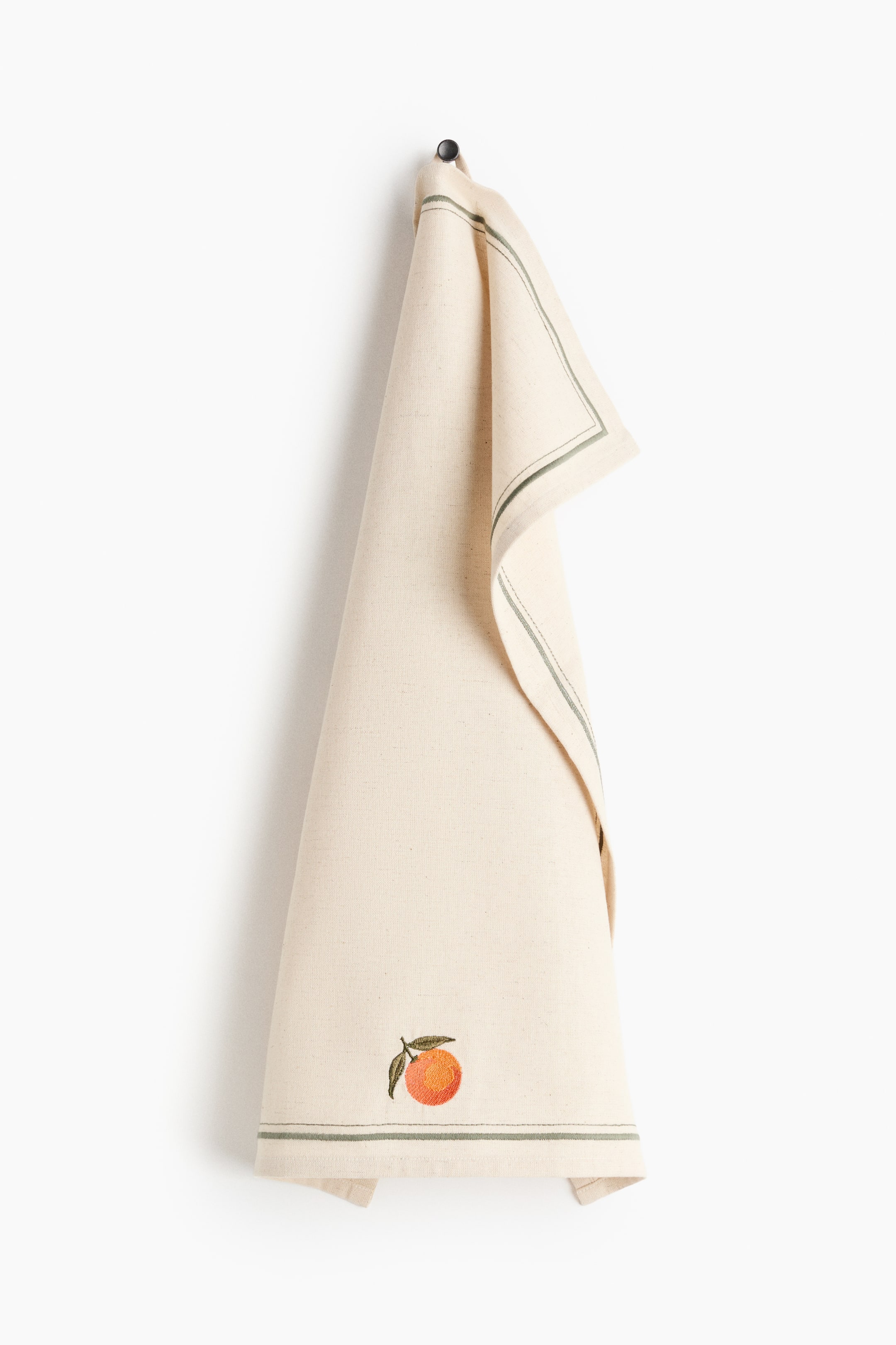 View larger image: A cream cotton-linen blend tea towel, folded lengthwise, hangs from a dark knob by a fabric loop on its upper short edge, featuring an embroidered orange with a leaf and parallel green stripes bordering the bottom and one vertical side.