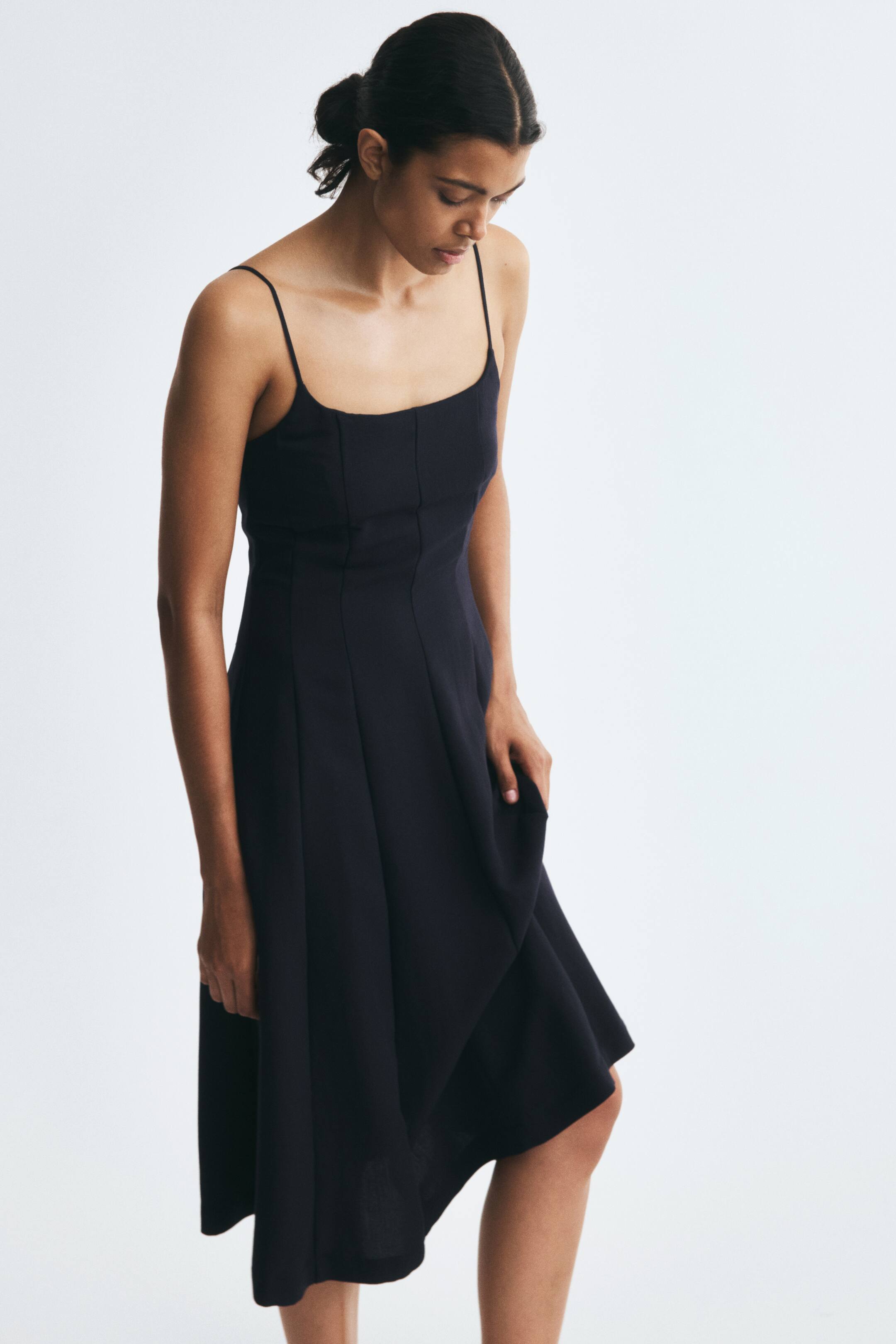 View larger image: A calf-length, black fitted dress featuring a scoop neckline, thin shoulder straps, and prominent vertical seams is being worn by the model. The model is holding a section of the flared A-line skirt while looking downward.