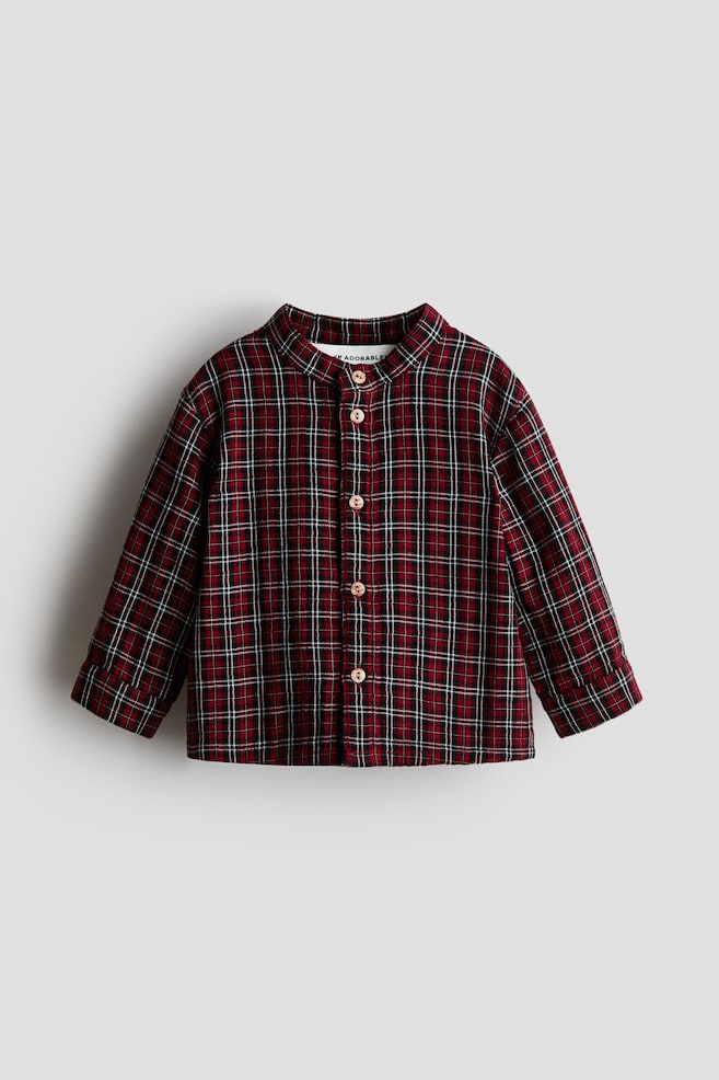 A plaid shirt, featuring a deep red, black, and white pattern, is presenting its front view. It is designed with a band collar and a front placket that is secured by four light-colored buttons, and its long sleeves are ending in simple hems. - 1