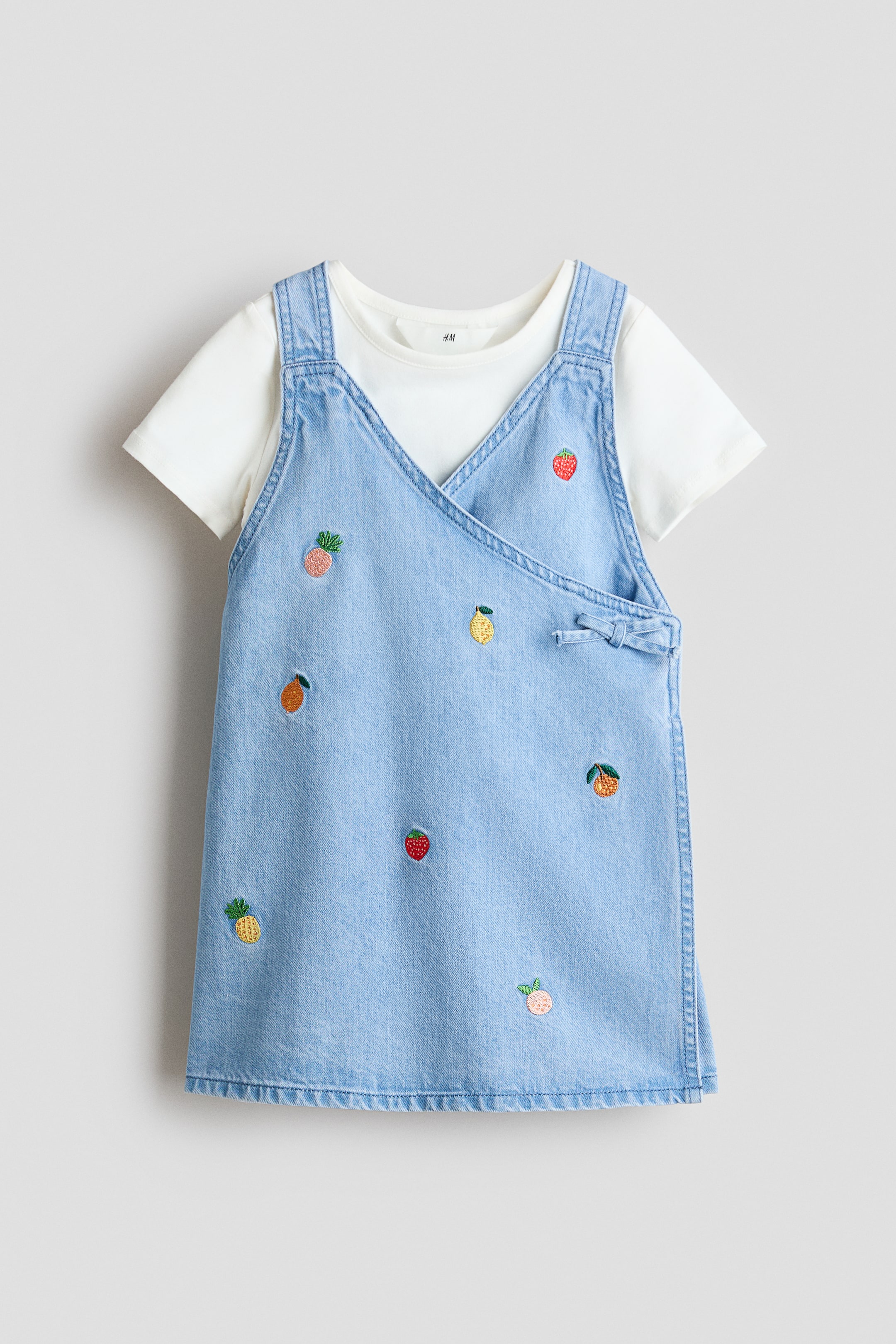 View larger image: A light blue denim dungaree dress is shown from the FRONT, layered over a white short-sleeved T-shirt. The denim dress features a V-neck wrap-style design with shoulder straps, a small bow detail on the right side, and is embellished with scattered embroidered fruit motifs.