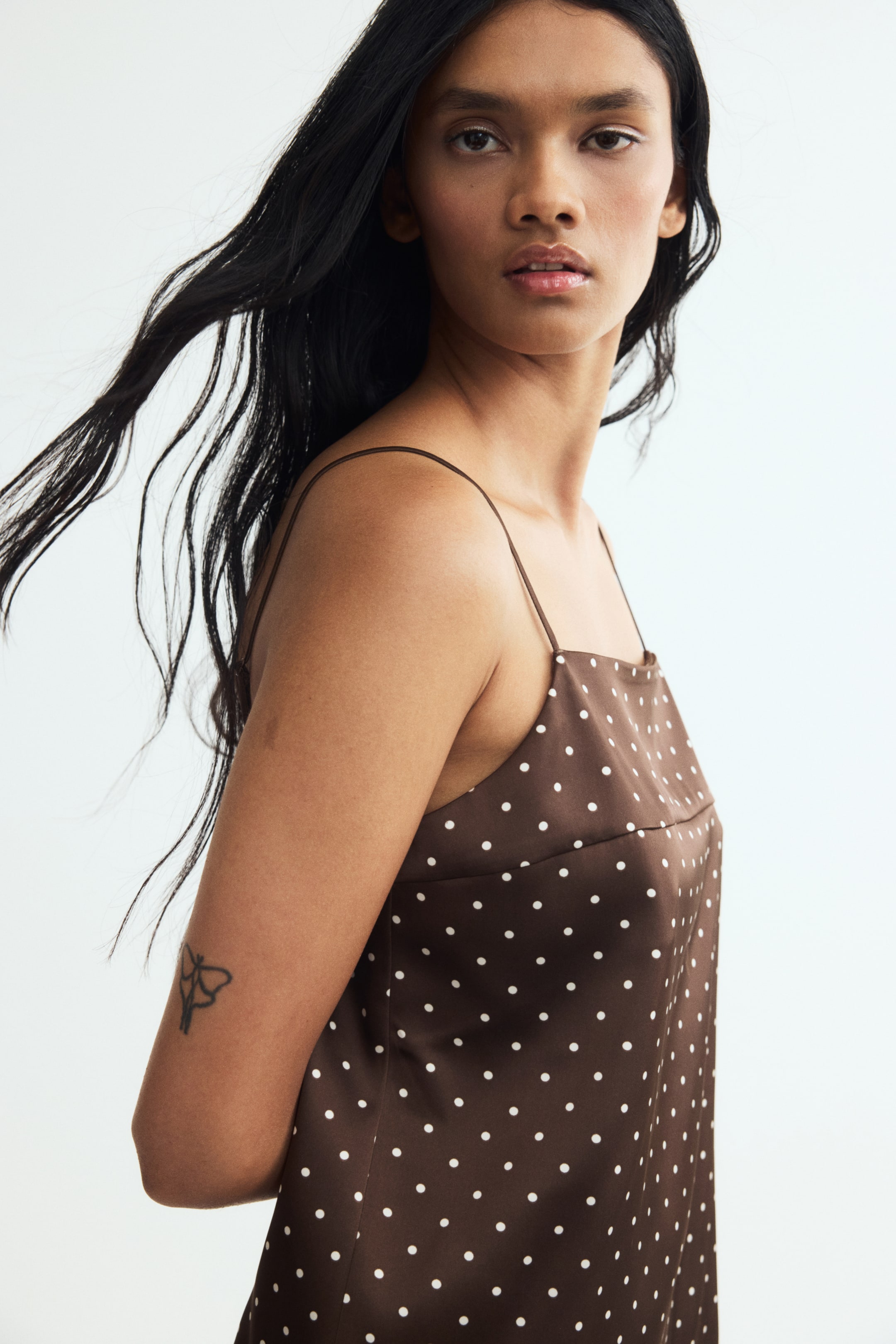 View larger image: The model is wearing a brown slip dress with a white polka-dot pattern, a square neckline, and thin spaghetti straps. The satin-like fabric has a smooth texture and a subtle sheen.