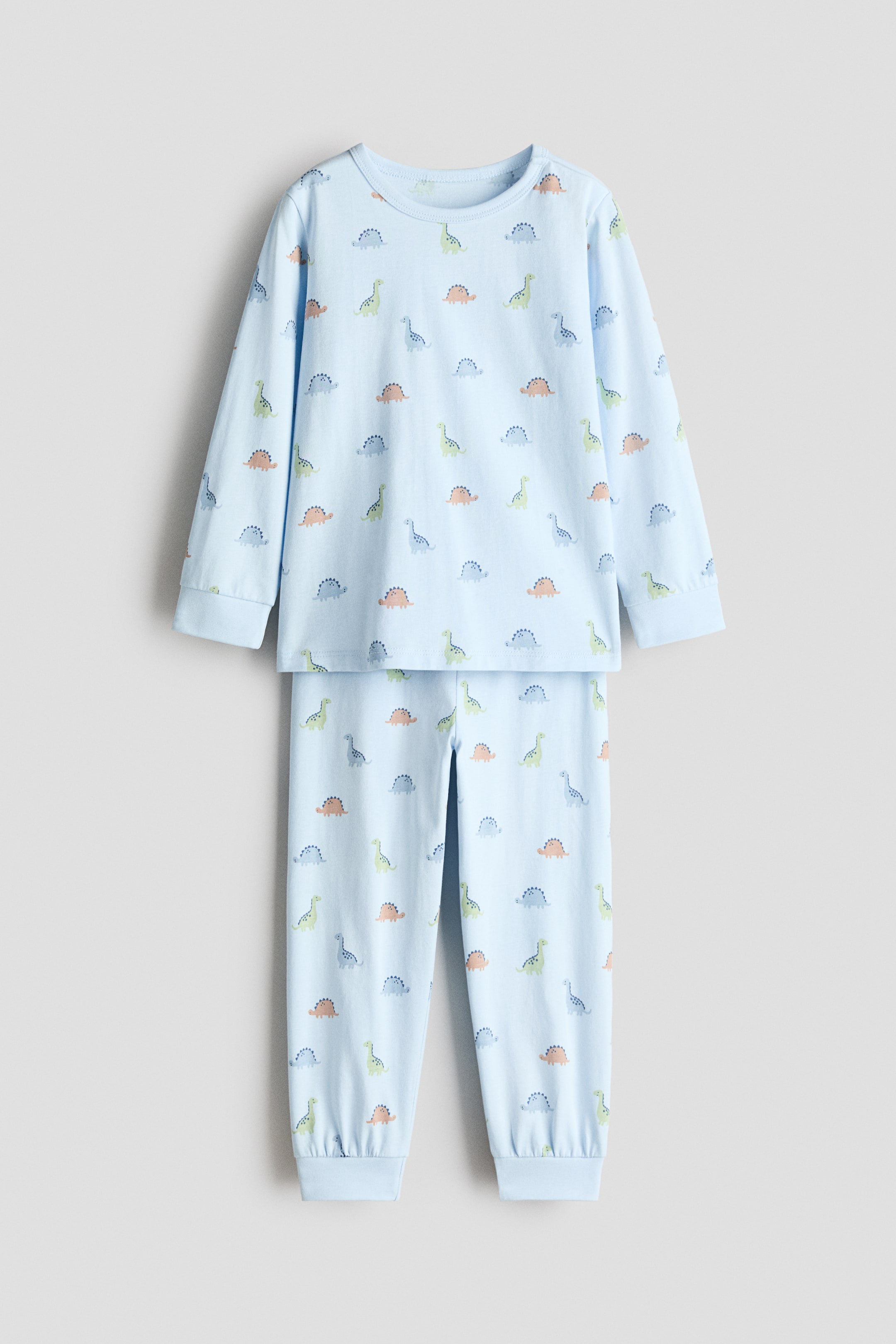 Agrandir l'image: Des pyjamas en coton imprimé sont posés à plat et face avant, composés d'un haut à manches longues bleu clair et d'un pantalon long assorti. Les deux pièces présentent un motif répétitif de petits dinosaures verts, orange et bleus. Le haut est conçu avec un col rond et des poignets côtelés, et le pantalon est doté de bas de jambes côtelés.