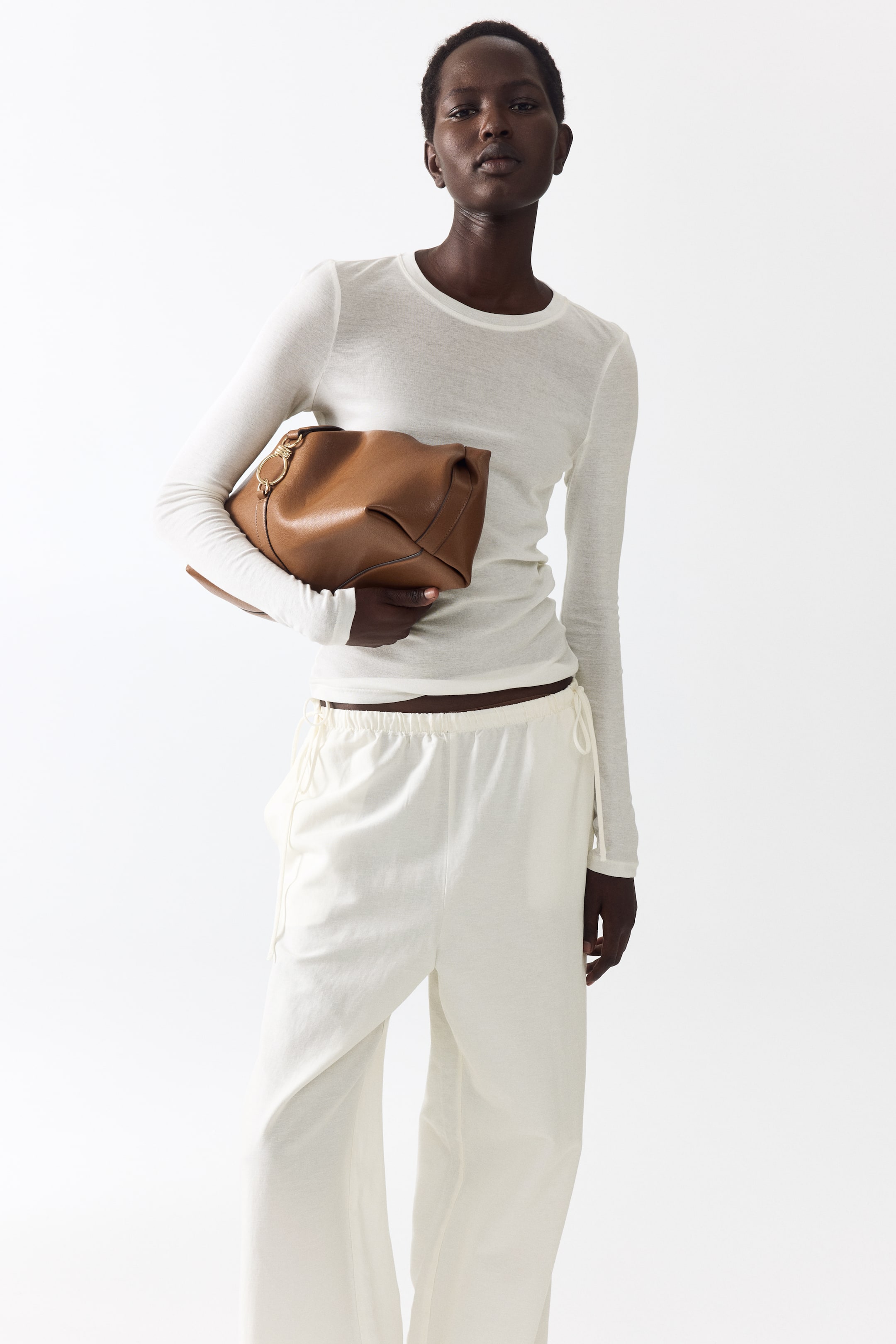 View larger image: A soft brown leather bag is being held under the arm, featuring a gold-toned circular hardware detail near its folded top and visible stitching along its edges.