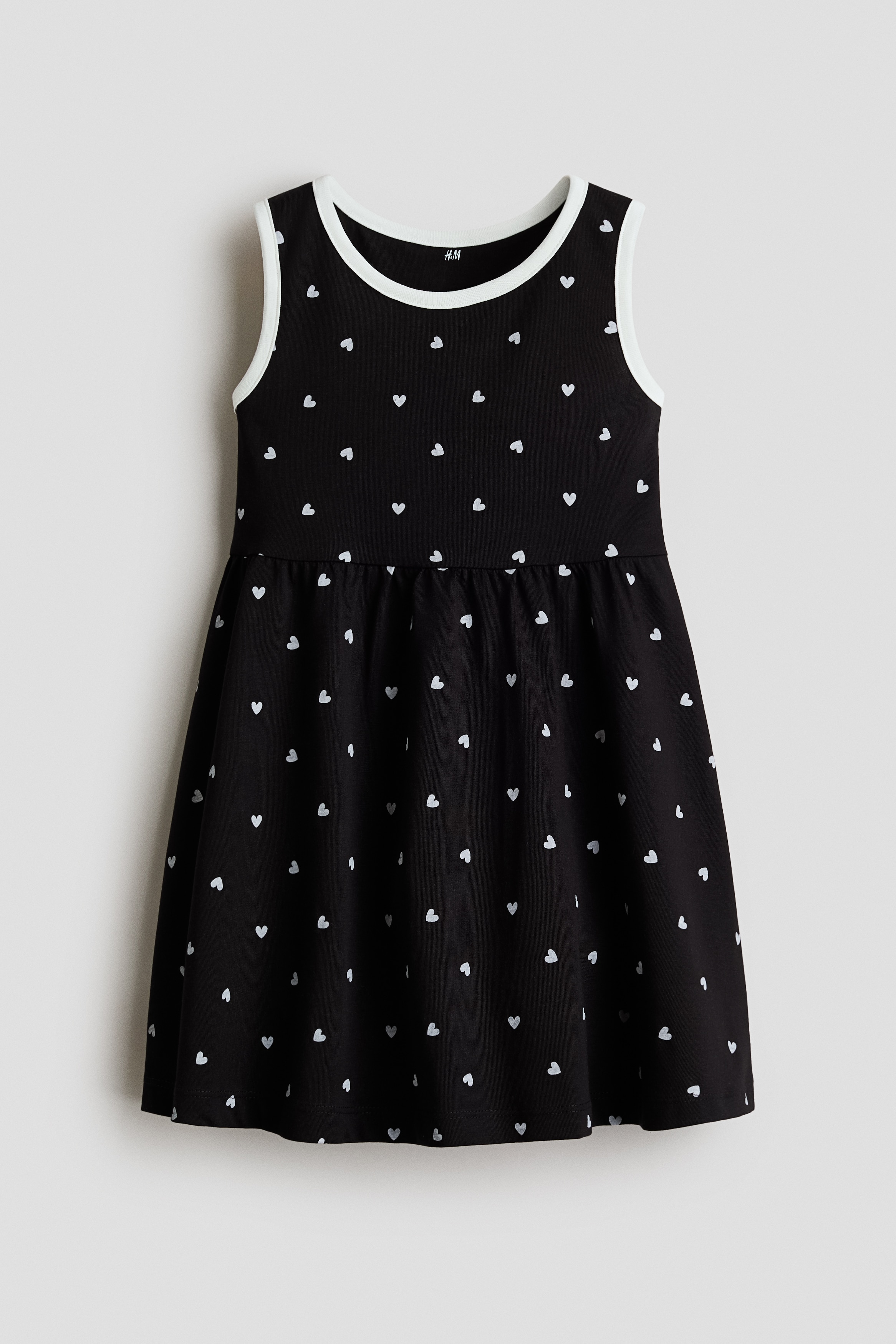 A sleeveless black dress featuring a small white heart pattern and white trim at the neckline and armholes is worn by the model. The dress has a gathered waistline and a relaxed fit.